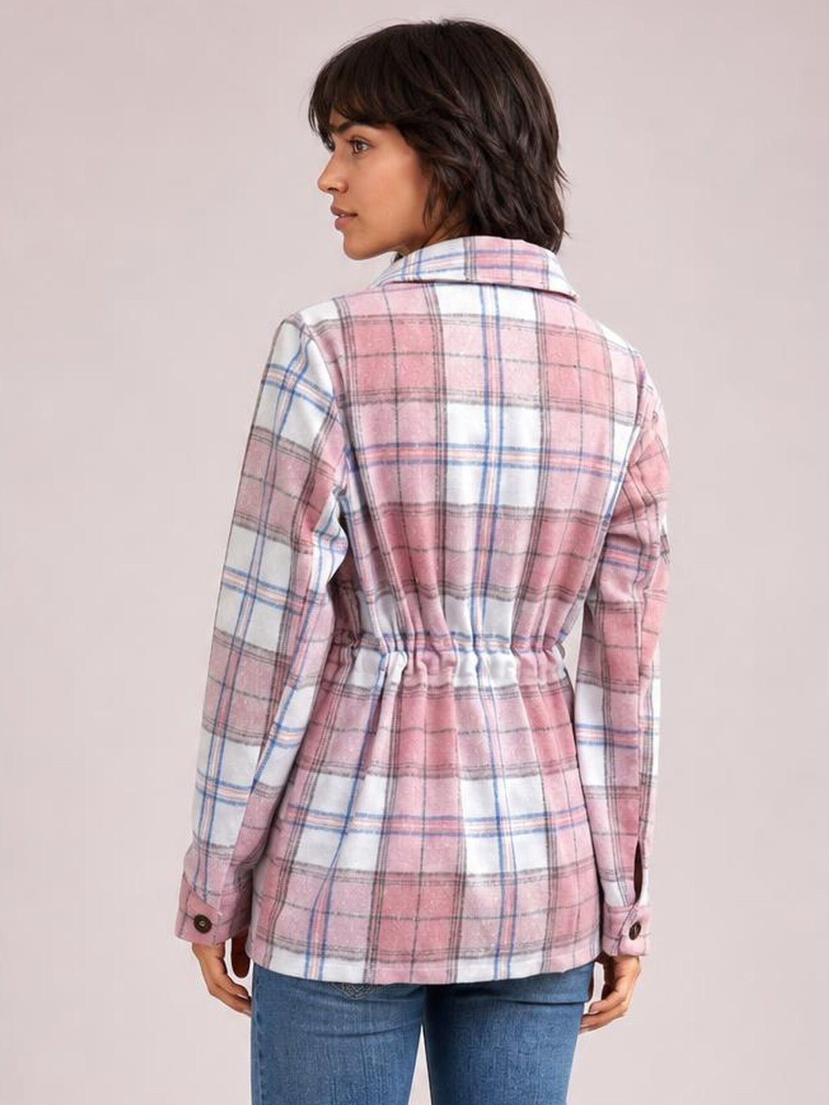 Model turned slightly to show the back of a pink, white, and blue plaid button-up jacket with long sleeves, cuff buttons, and a drawstring waist.