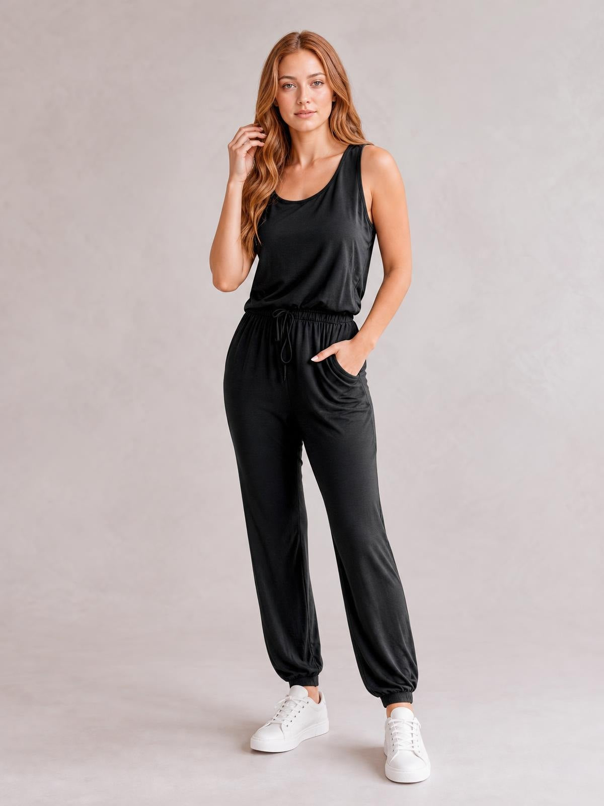 A model poses in a black sleeveless round-neck jumpsuit with a drawstring waist, relaxed jogger silhouette, tapered legs, and side pockets.