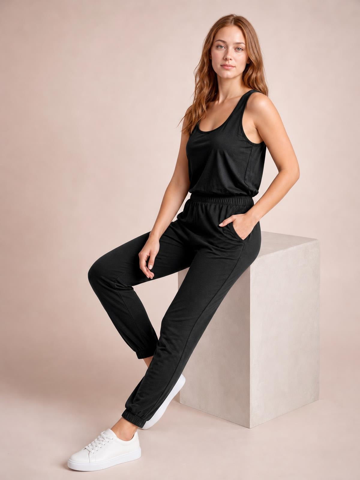 Model seated in a black sleeveless round-neck jumpsuit with a relaxed tapered silhouette, elastic waist, drawstring detail, and practical side pockets.