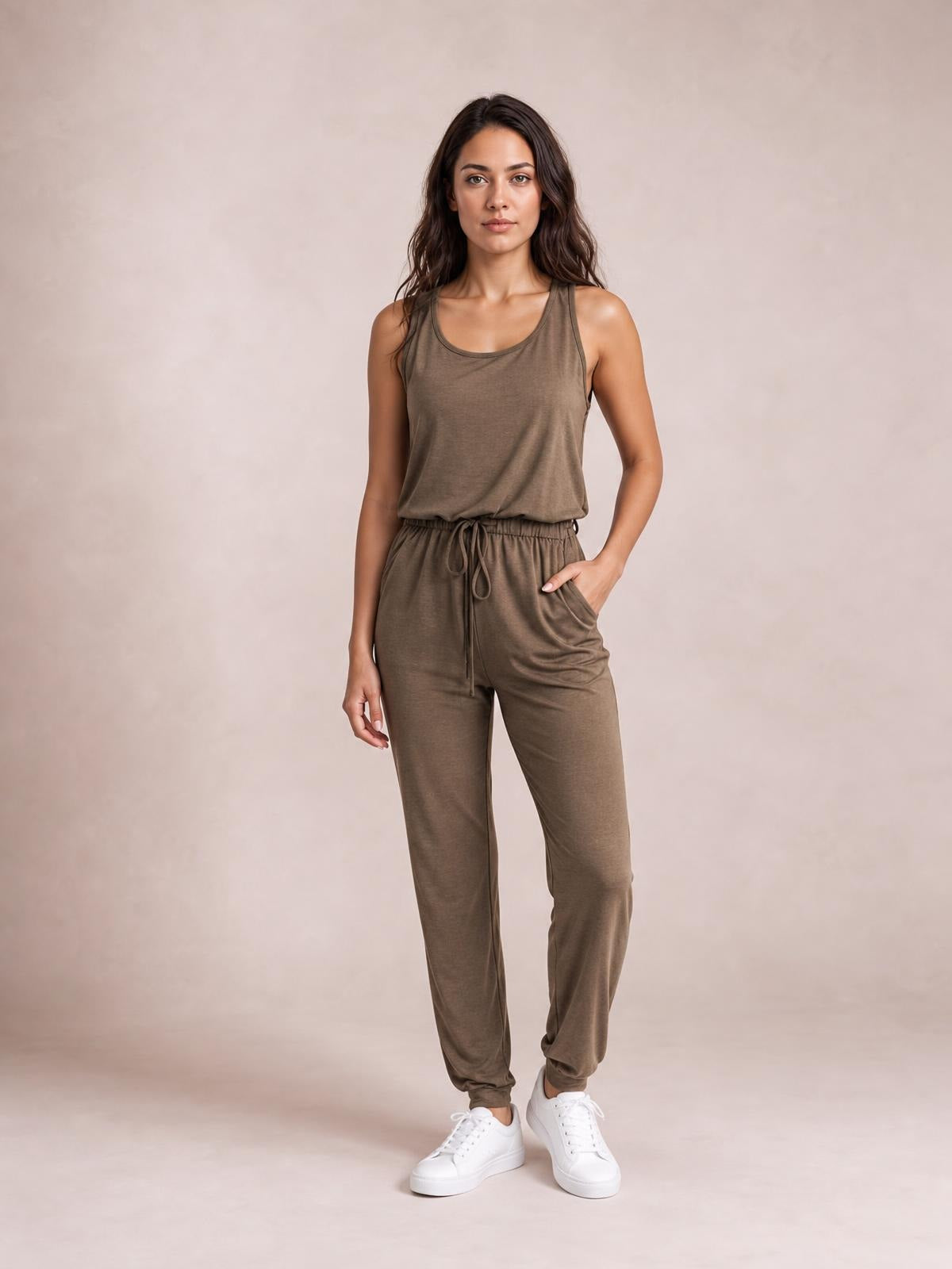 Model wears a taupe sleeveless round-neck jumpsuit with a relaxed silhouette, drawstring waist, tapered legs, and side pockets, standing with one hand in a pocket.