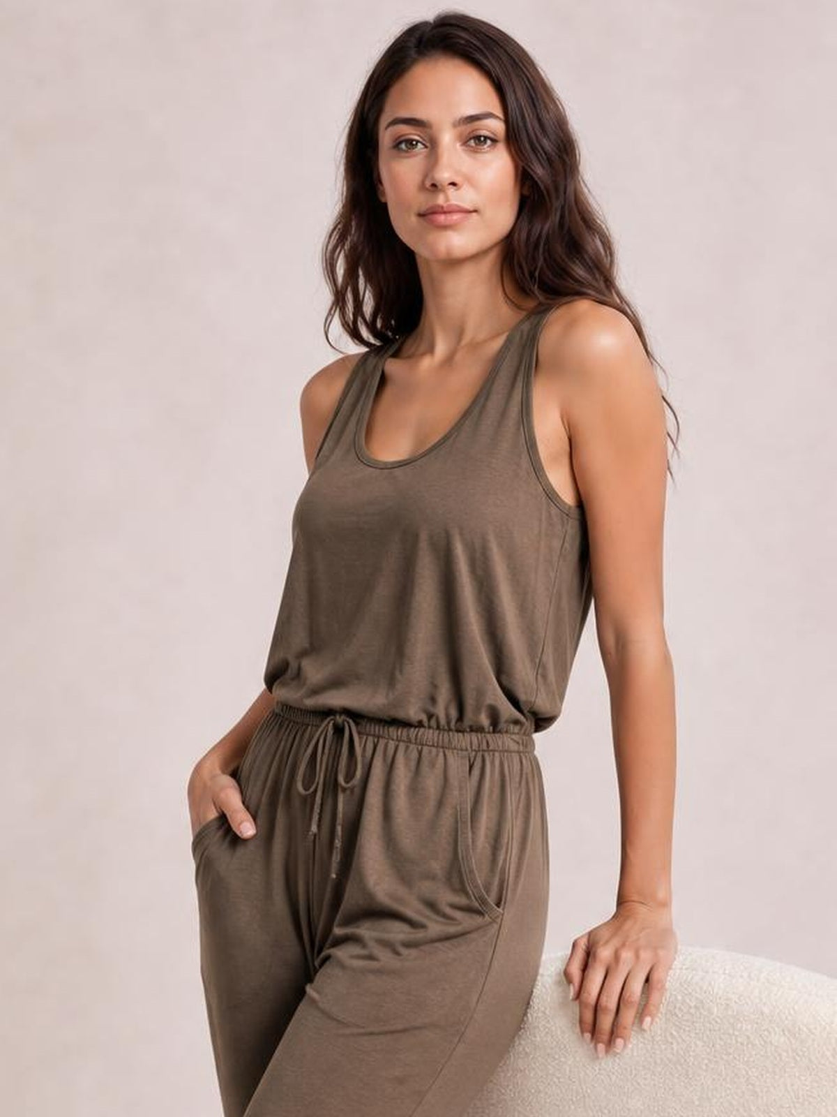 Model wearing a brown sleeveless jumpsuit with a round neck, drawstring waist, relaxed silhouette, and roomy side pockets, posing with one hand inside a pocket.