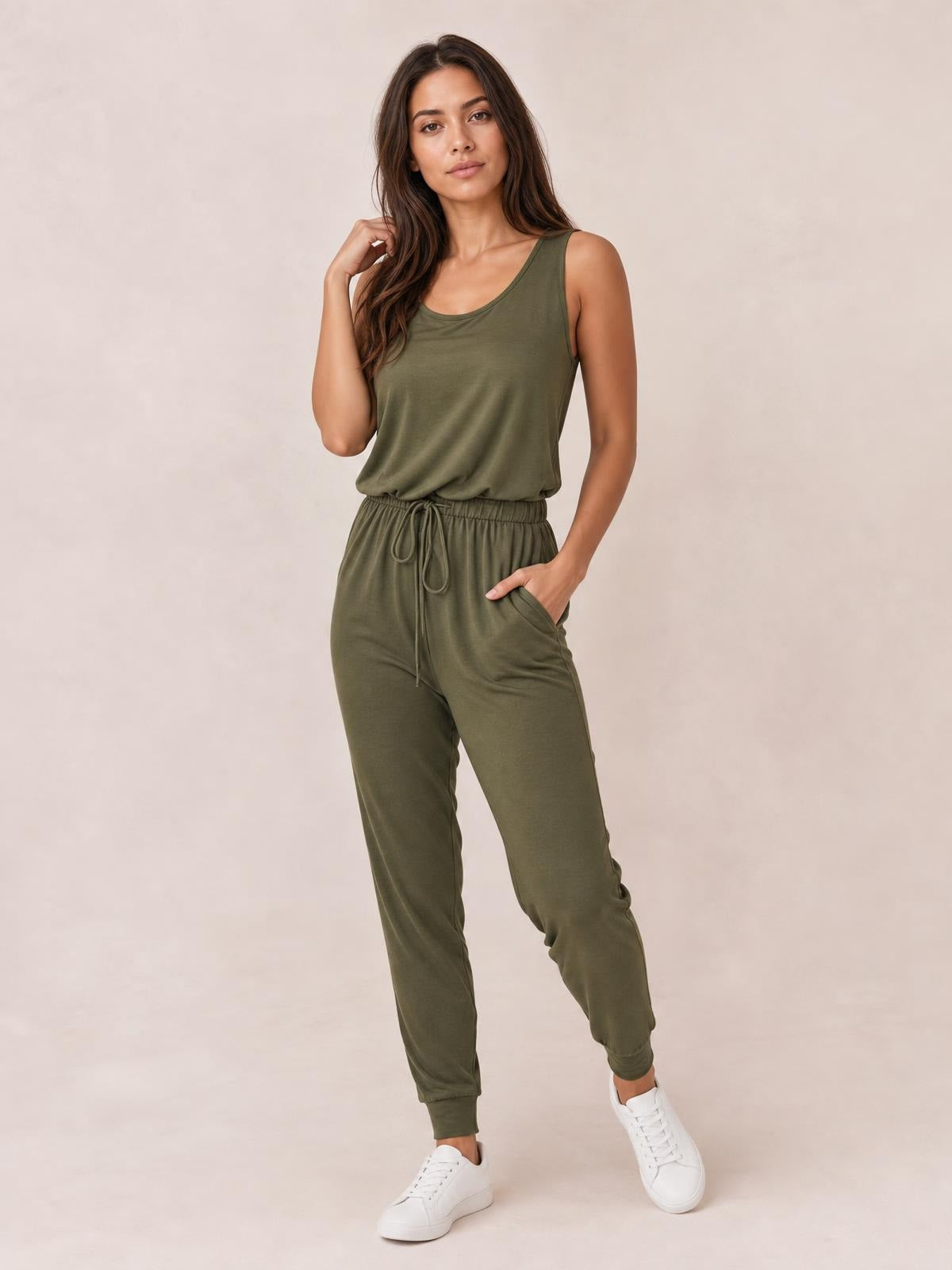 A model poses in an olive-green sleeveless round-neck jumpsuit with a drawstring waist, side pockets, and relaxed tapered legs.