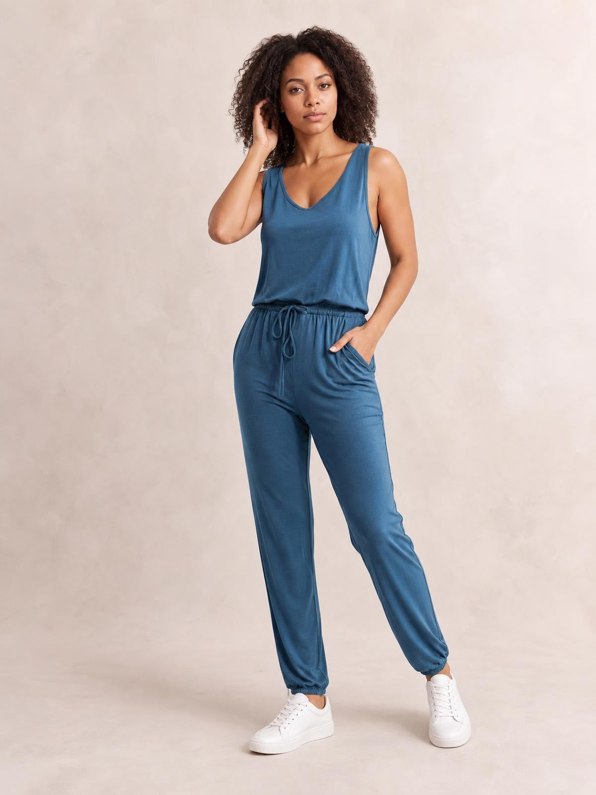 Model poses in a blue sleeveless round-neck jumpsuit with a drawstring waist, relaxed tapered legs, and side pockets.