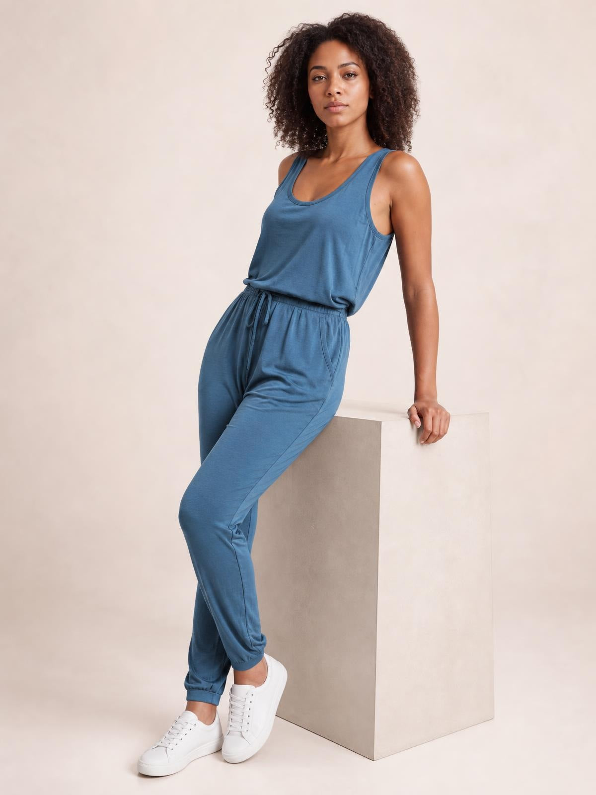 Model poses in a blue drawstring round-neck sleeveless jumpsuit with a relaxed tapered silhouette, elastic cuffs, and side pockets.