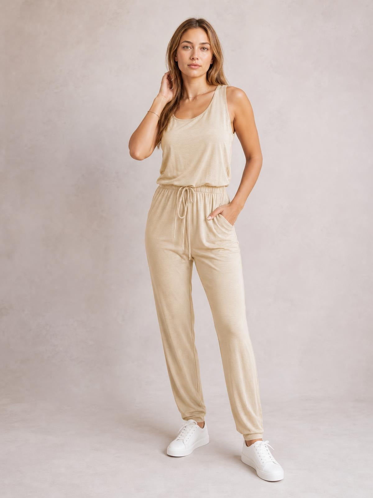 Model wears a beige sleeveless jumpsuit with a round neck, drawstring waist, relaxed tapered legs, and side pockets, posing with one hand tucked inside.