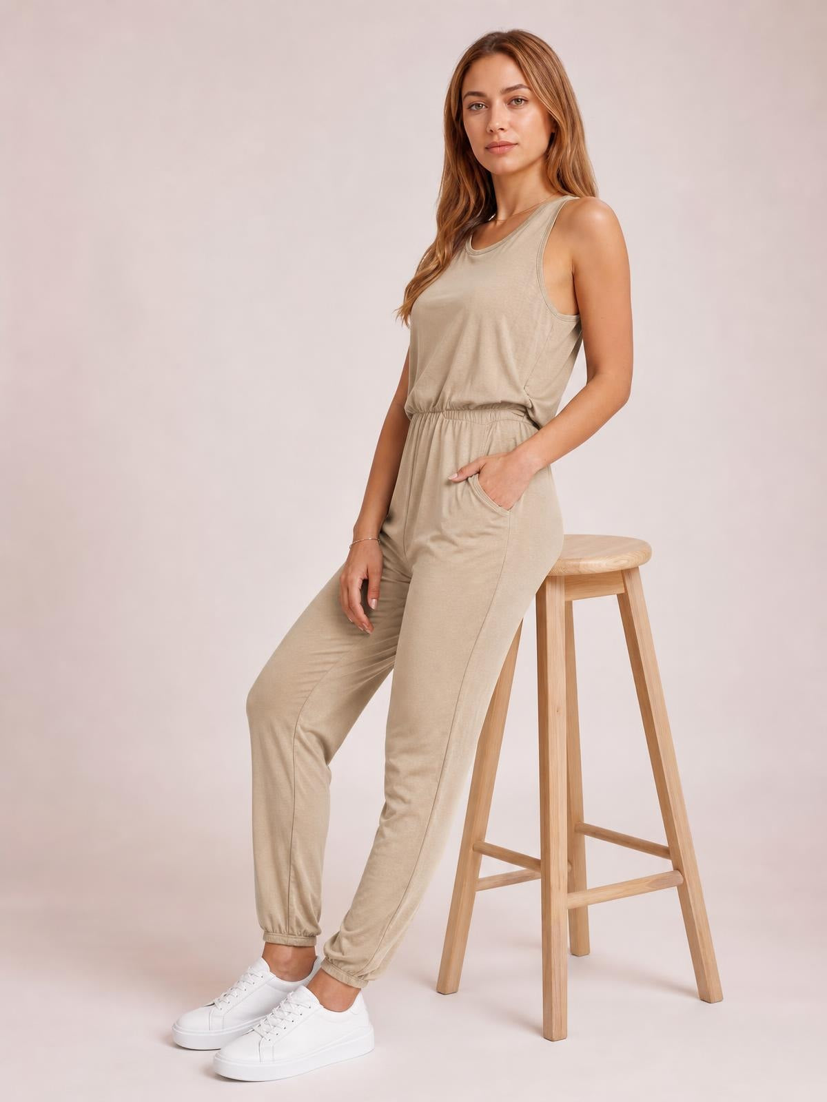 Model poses beside a stool in a beige sleeveless jumpsuit with a round neck, drawstring waist, tapered legs, and a visible side pocket.