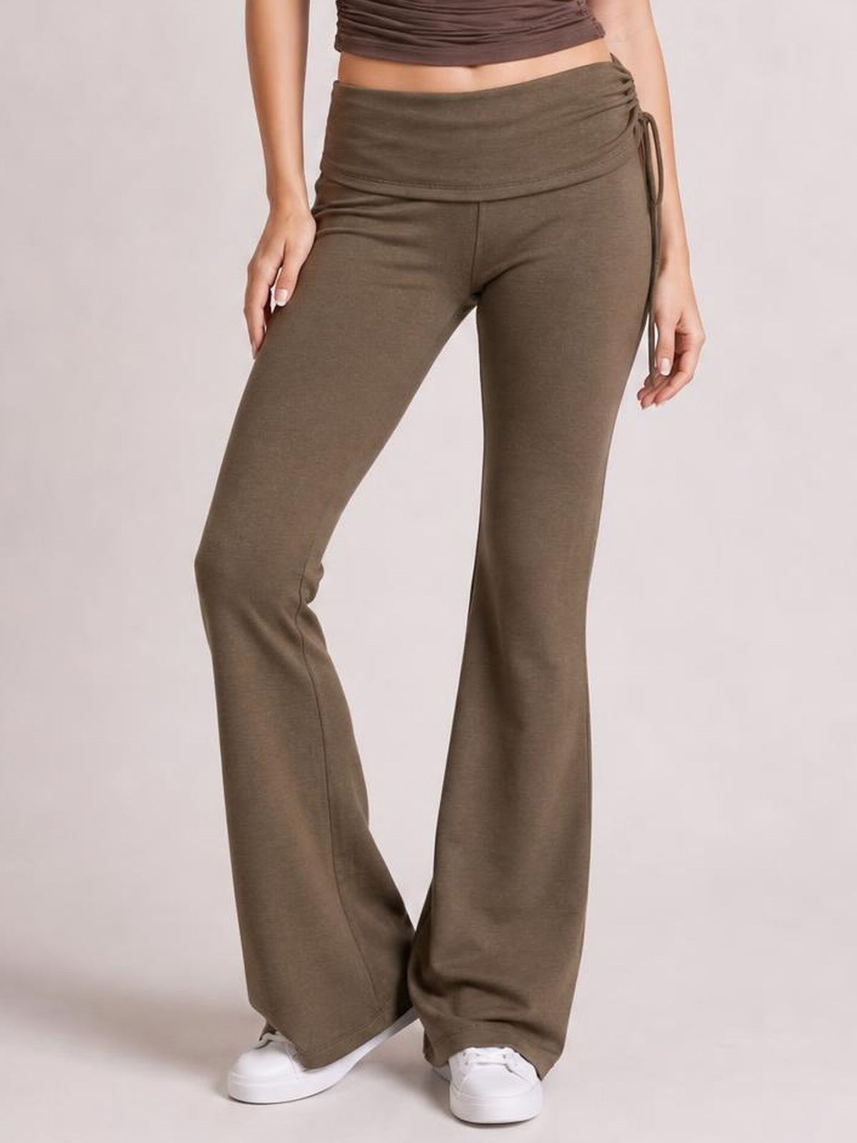 A model stands with one knee bent, wearing brown drawstring ruched flare pants with a fitted silhouette through the hips and wide, flowing hems.