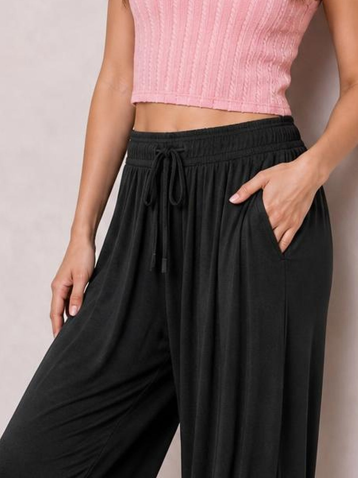 Model wearing black drawstring ruched pants with an elastic waistband, wide-leg silhouette, and flowing gathered construction, one hand in a pocket.
