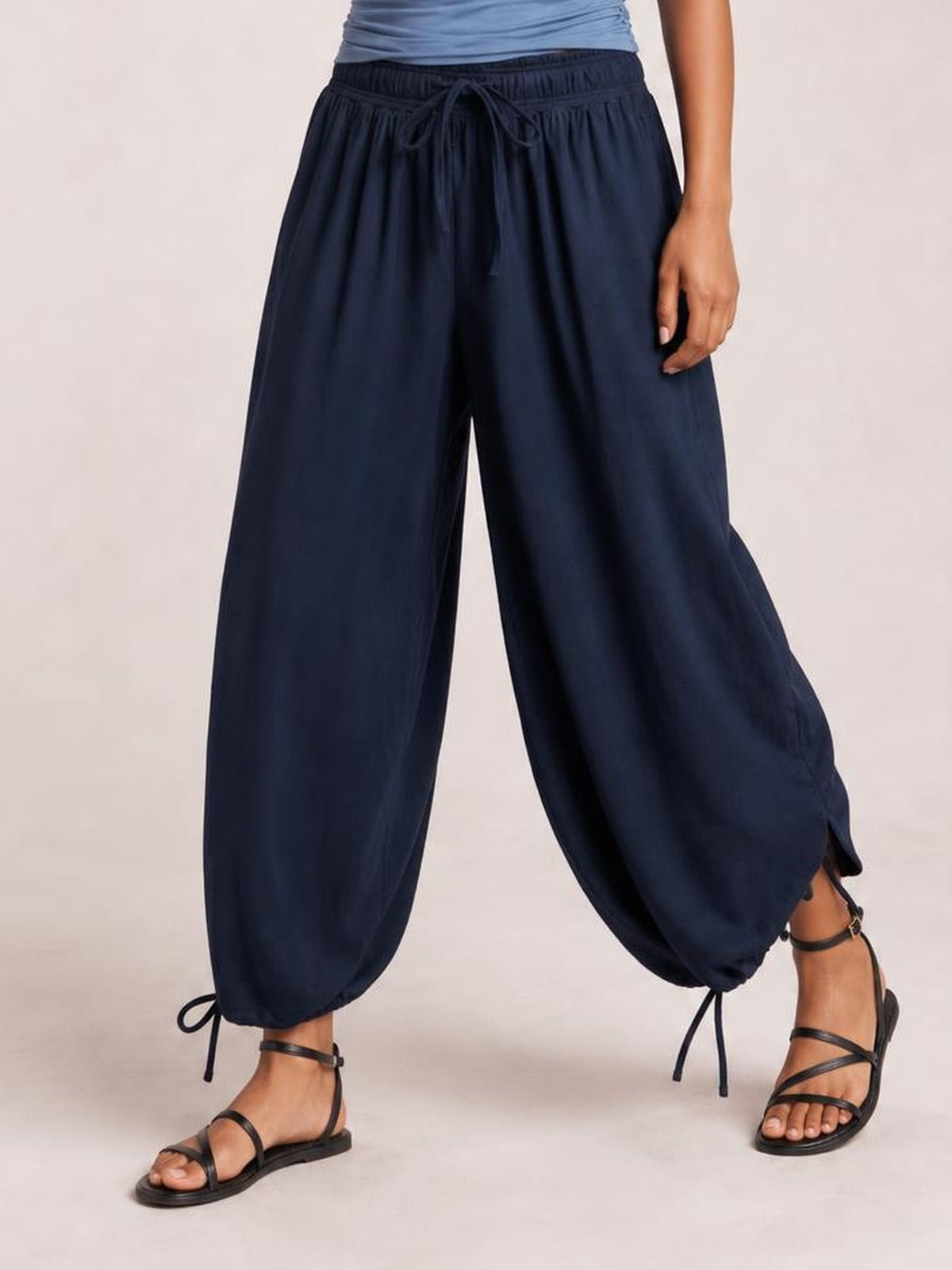 A model wears navy drawstring ruched pants with a gathered elastic waist, relaxed wide-leg silhouette, and adjustable ankle ties while standing in sandals.