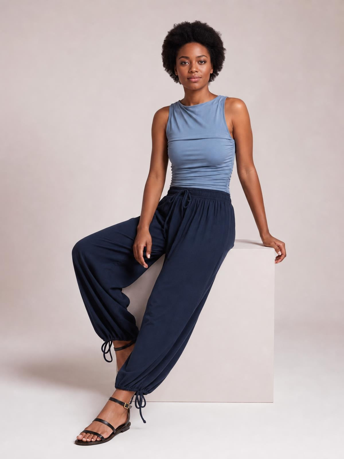 A seated model wears navy drawstring ruched pants with a relaxed silhouette, elastic waist, gathered ankle cuffs, and adjustable ties.