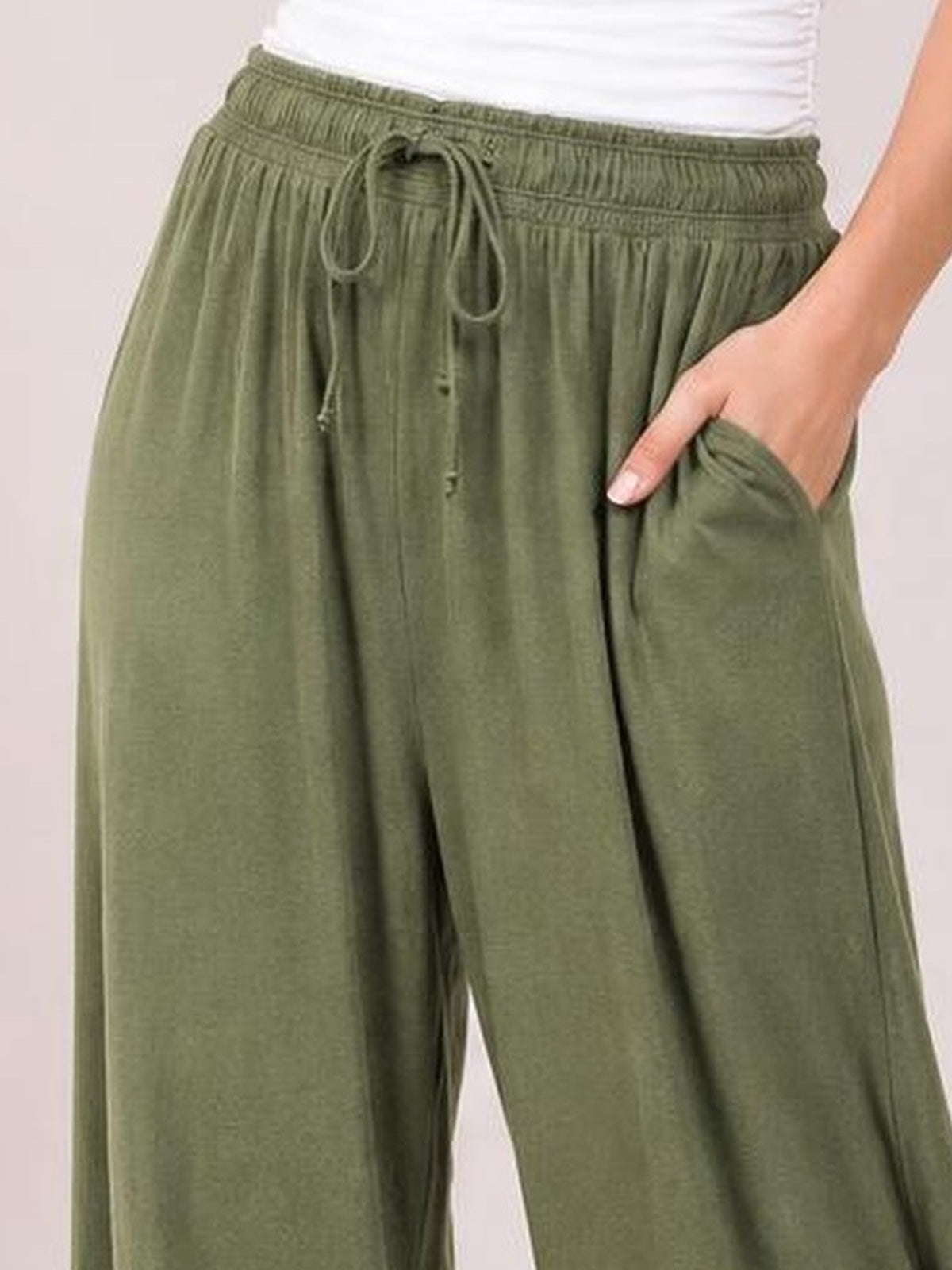 Cropped model wears olive green drawstring ruched pants with an elastic waist, wide relaxed legs, vertical gathers, and side pockets, posing with one hand tucked in.