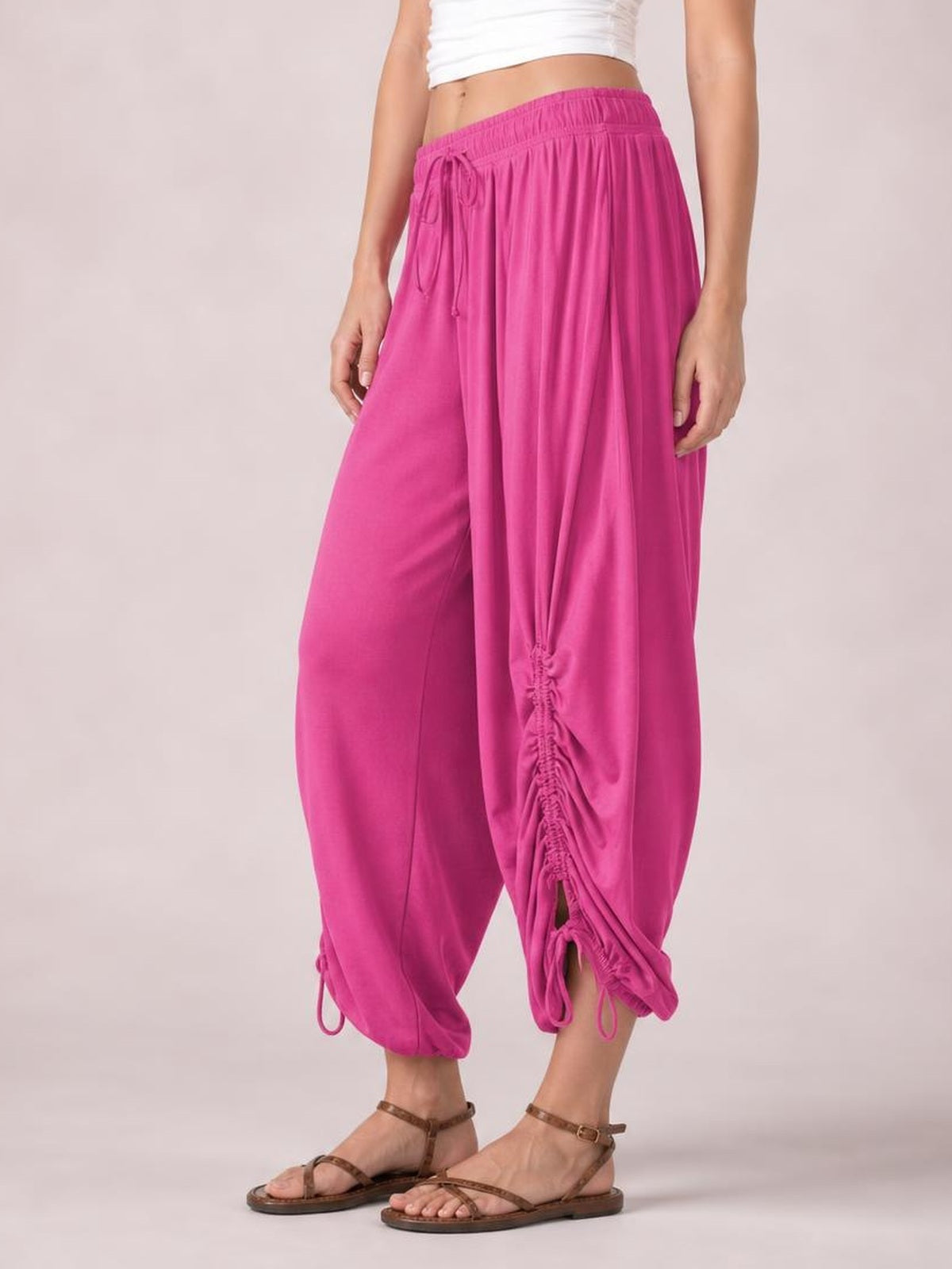 Model wears bright pink drawstring ruched pants with a relaxed cropped silhouette, elastic waistband, gathered lower-leg detailing, and adjustable ankle ties.