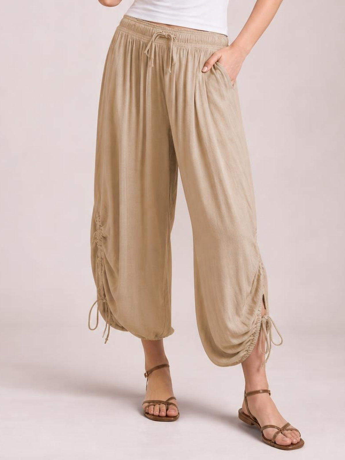 Model wears beige drawstring ruched pants with a relaxed wide-leg silhouette, gathered ankle details, and side pockets while posing with one hand tucked in a pocket.