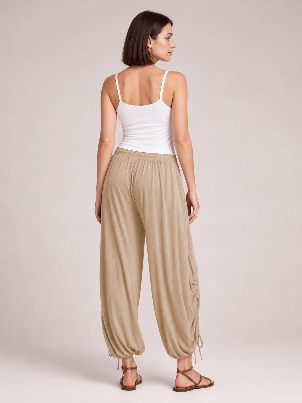Model in a back-facing pose wears beige drawstring ruched pants with a relaxed wide-leg silhouette, elastic waist, and gathered ankle hems with ties.