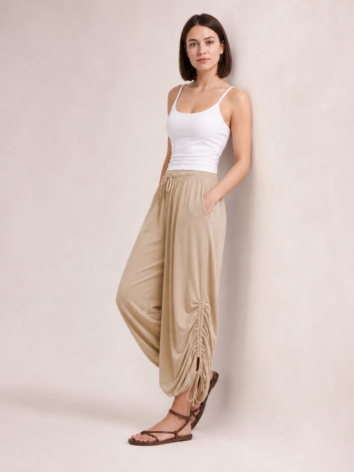 A model poses in beige drawstring ruched pants with an elastic waist, wide relaxed legs, side pockets, and gathered ankle detailing, one hand tucked in a pocket.