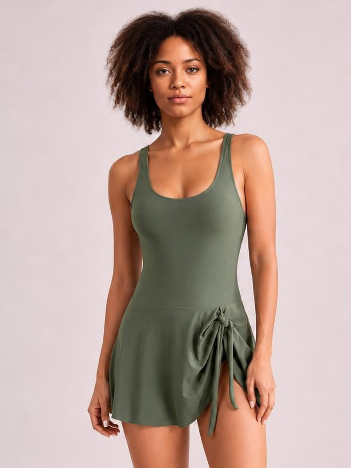 Model stands in an olive-green one-piece swimsuit with a scoop neck, wide straps, fitted silhouette, and a visible drawstring waist tie.