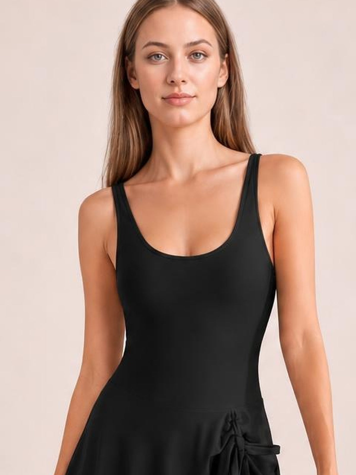 A model stands facing forward in a black drawstring scoop-neck one-piece swimsuit with wide straps, a fitted stretchy silhouette, and side waist tie.