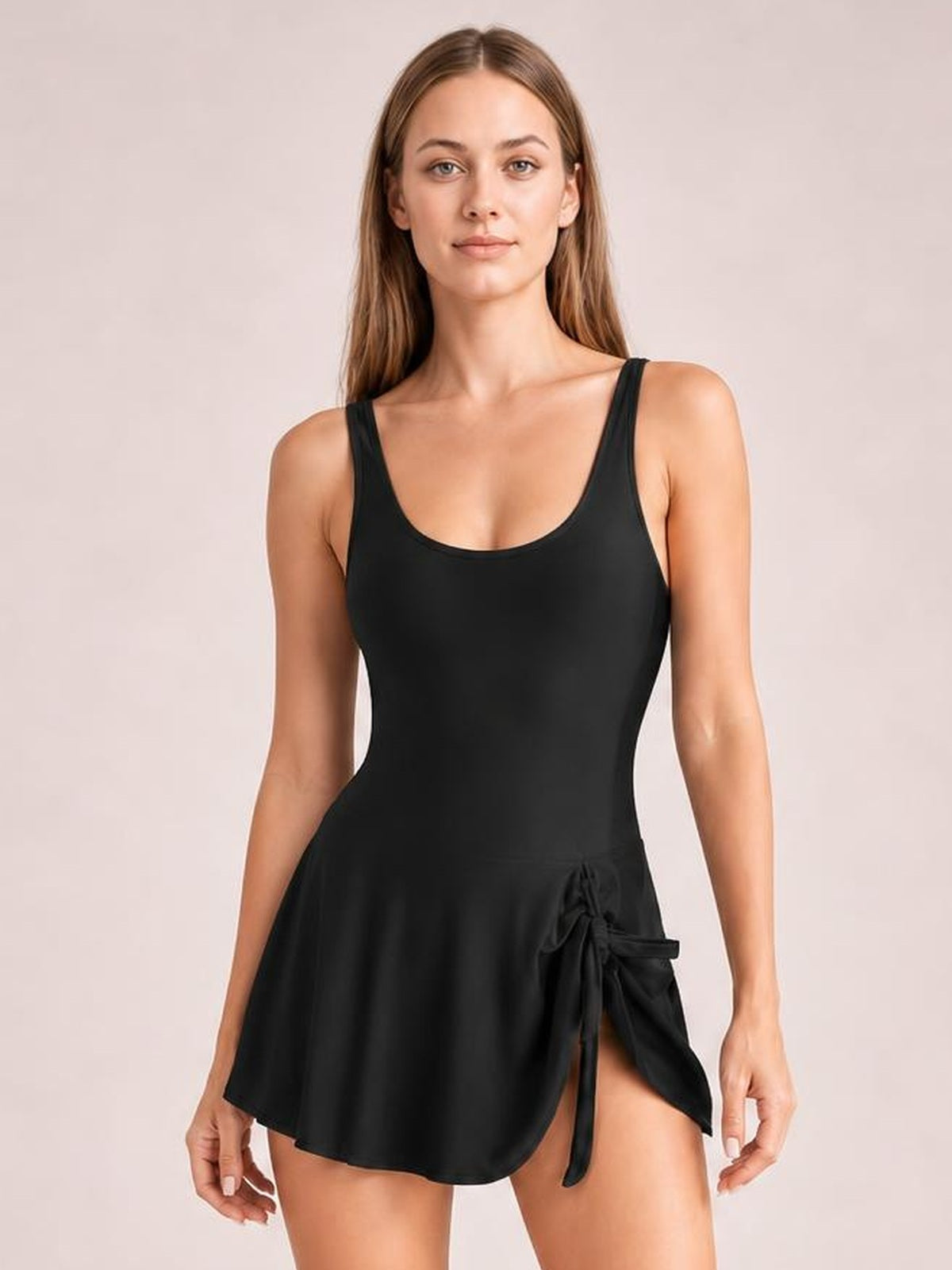 A model wears a black drawstring scoop neck one-piece swimsuit with wide straps, a fitted bodice, and a flared swim-dress silhouette with side tie.