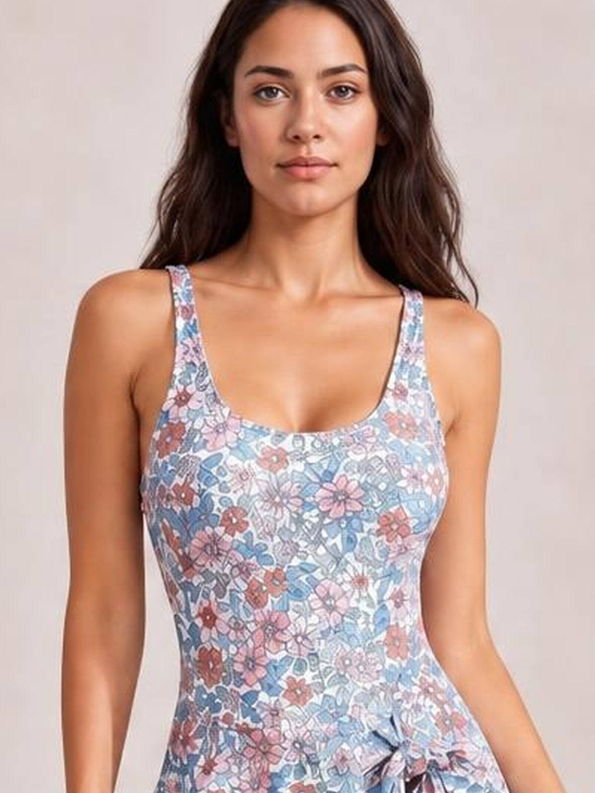 A model faces forward in a pastel blue and pink floral one-piece swimsuit with a scoop neck, wide straps, fitted silhouette, and front drawstring tie.