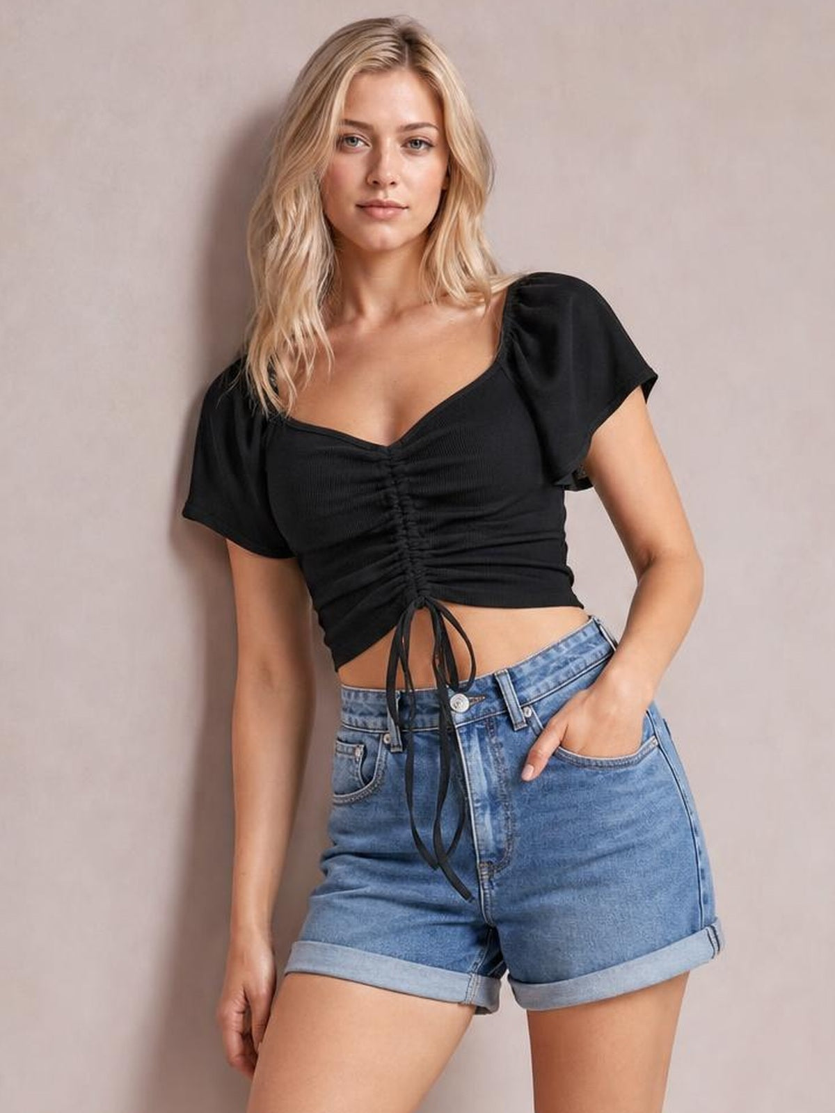 Model wears a black drawstring short sleeve crop top with a ruched front and fitted silhouette, posing with one hand in her denim shorts pocket.