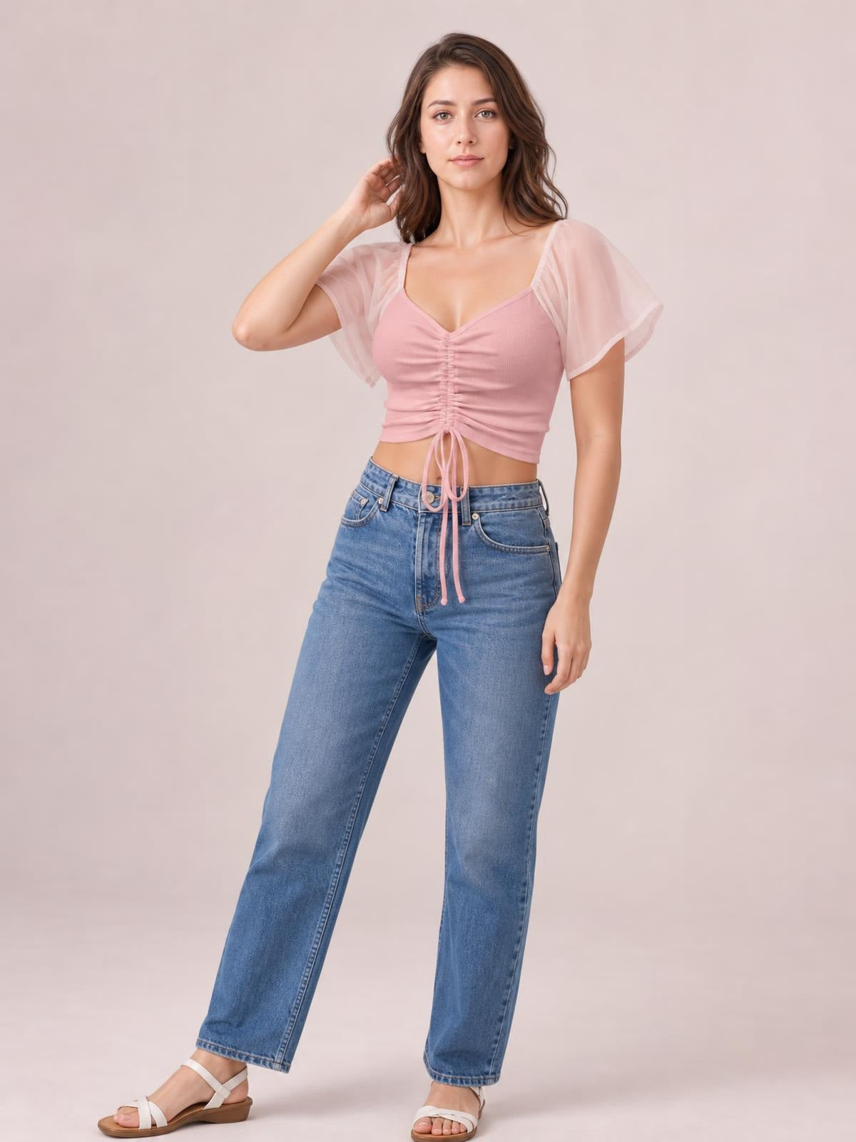 A model poses in a pink short sleeve crop top with a fitted silhouette, ruched drawstring front, and long ties over high-waisted jeans.