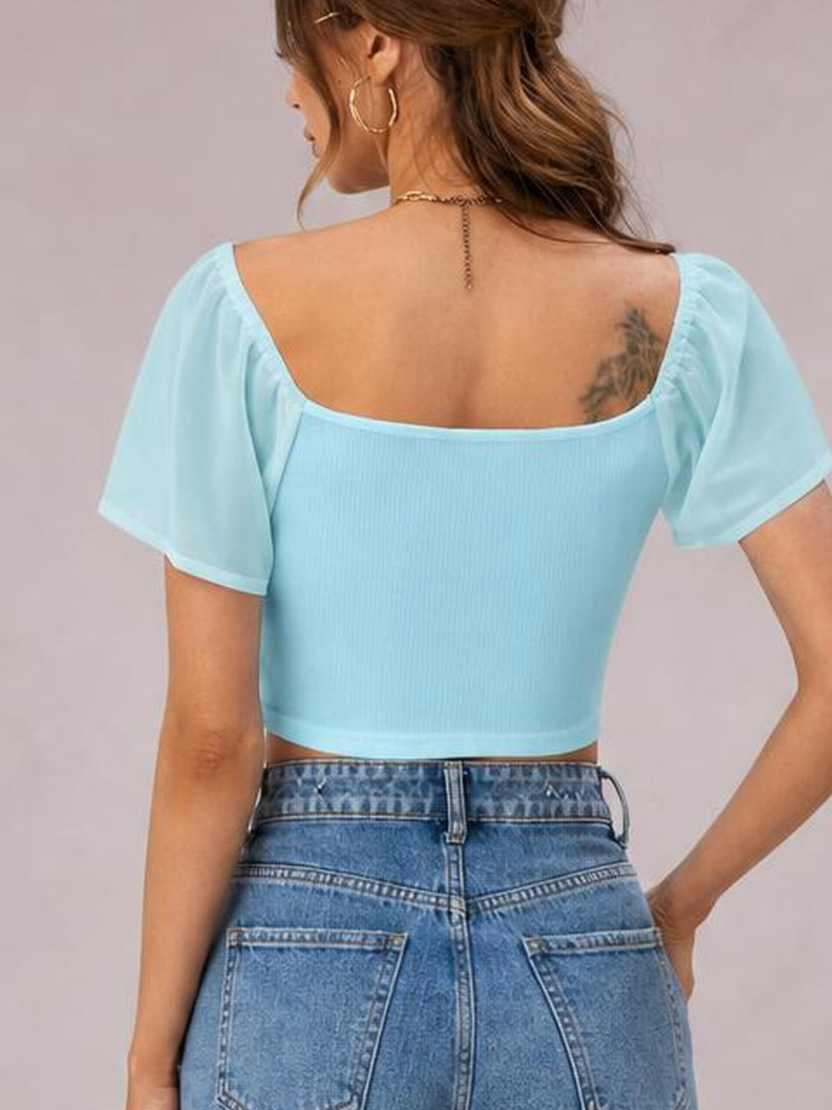 A model poses with her back to the camera in a light blue short sleeve crop top with a wide square back neckline and fitted ribbed silhouette.