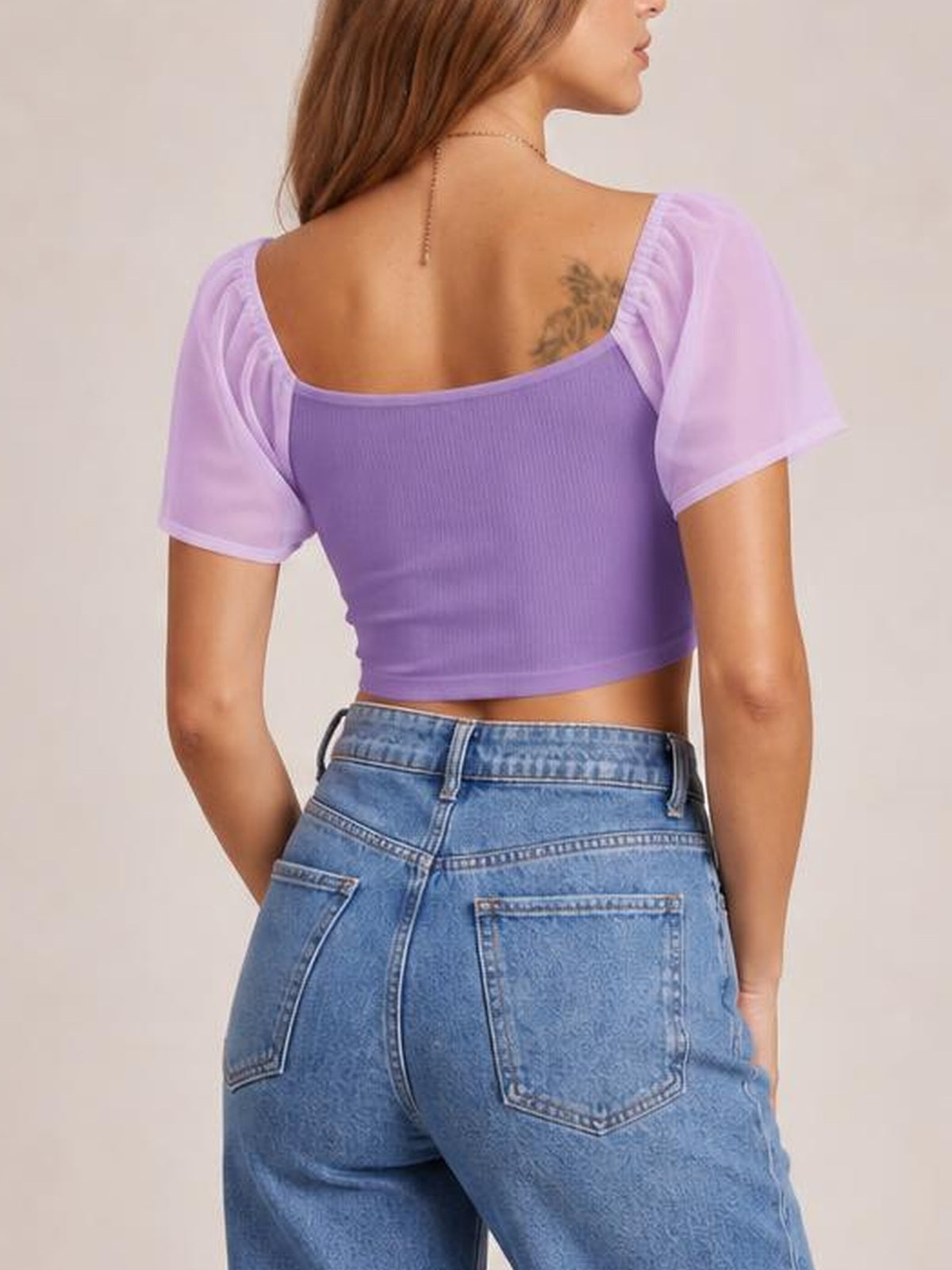 A model poses from the back in a lavender drawstring crop top with a fitted ribbed bodice, square neckline, and gathered short sleeves.