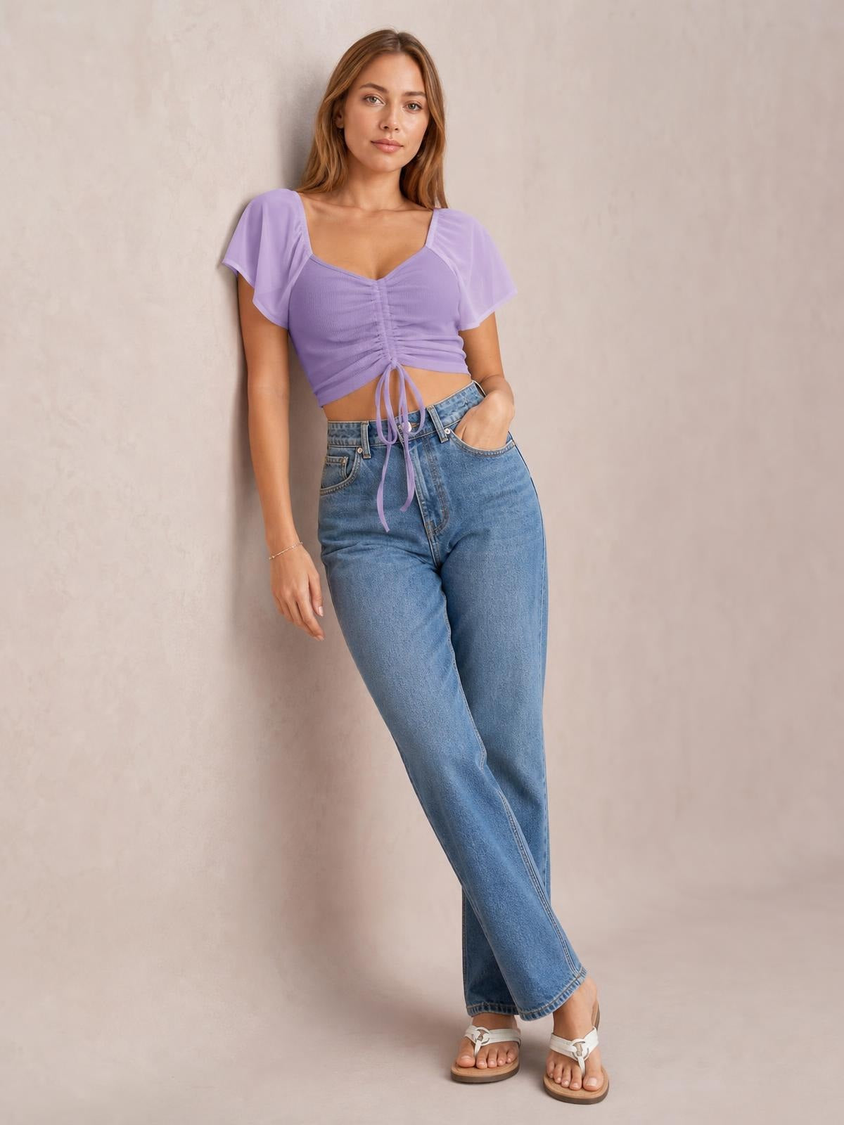 Model poses in a lavender short-sleeve crop top with ruched drawstring front and long ties, paired with relaxed blue jeans.