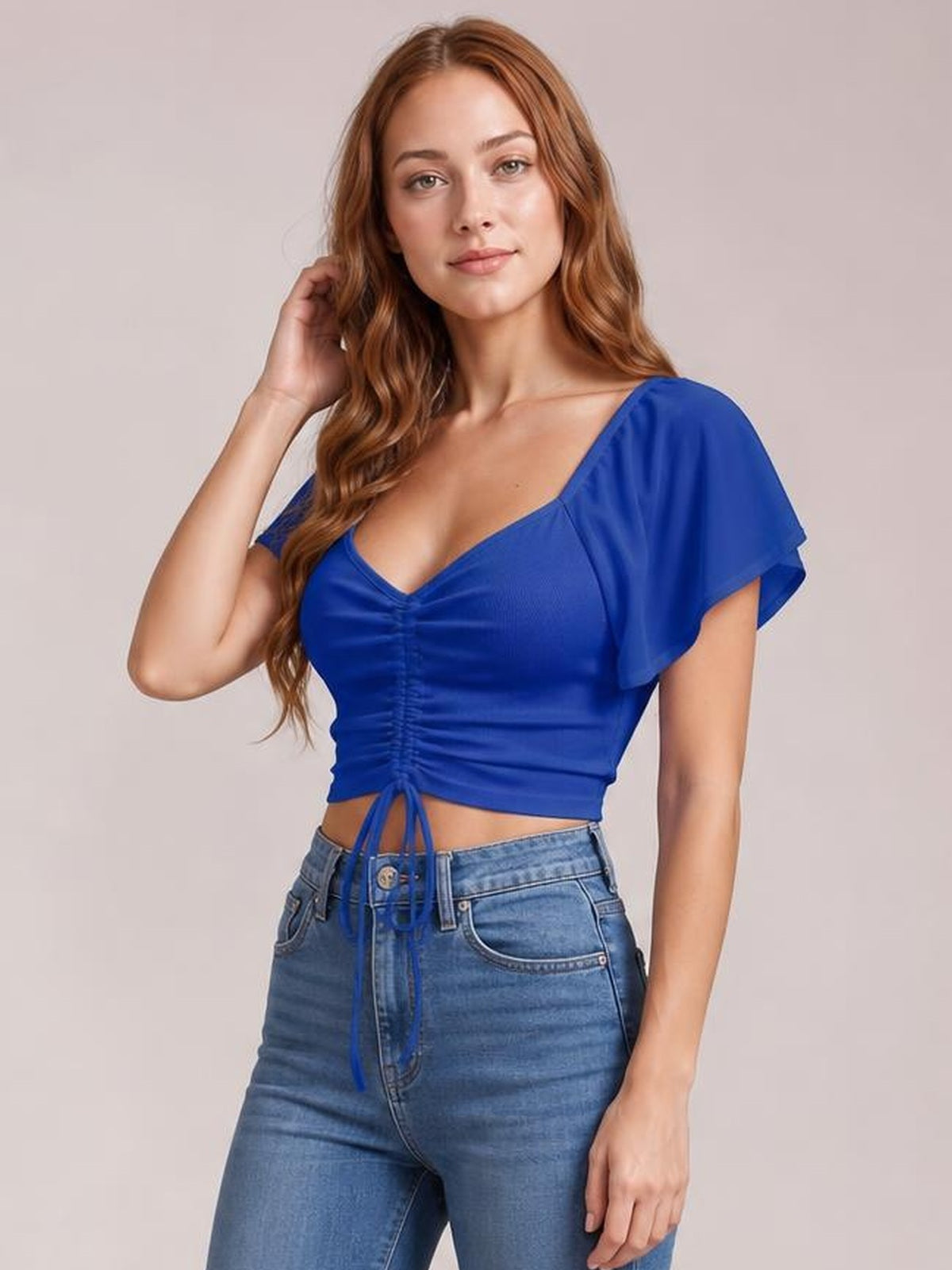 Model poses with one hand in her hair wearing a royal blue short-sleeve crop top with a ruched front, drawstring hem, and flared sleeves.