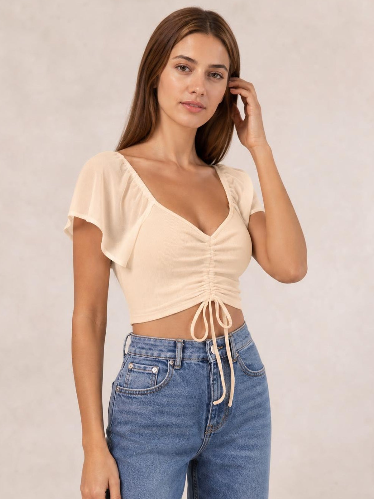 Model poses in a cream drawstring short sleeve crop top with a ruched front, V-neckline, relaxed sleeves, and adjustable tie hem.