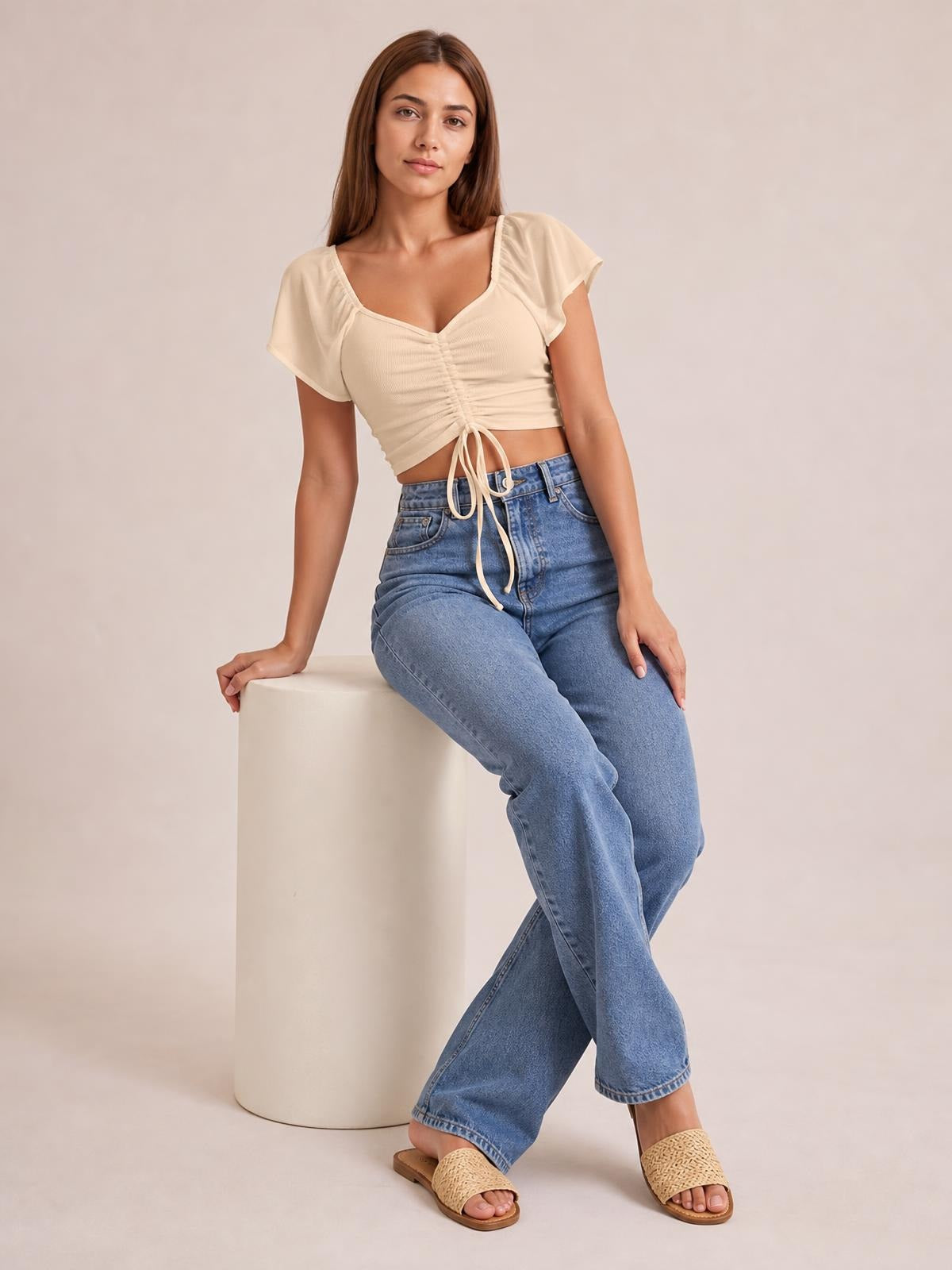 Model seated in a cream drawstring short sleeve crop top with a ruched front, tie detail, square neckline, and fitted cropped silhouette.