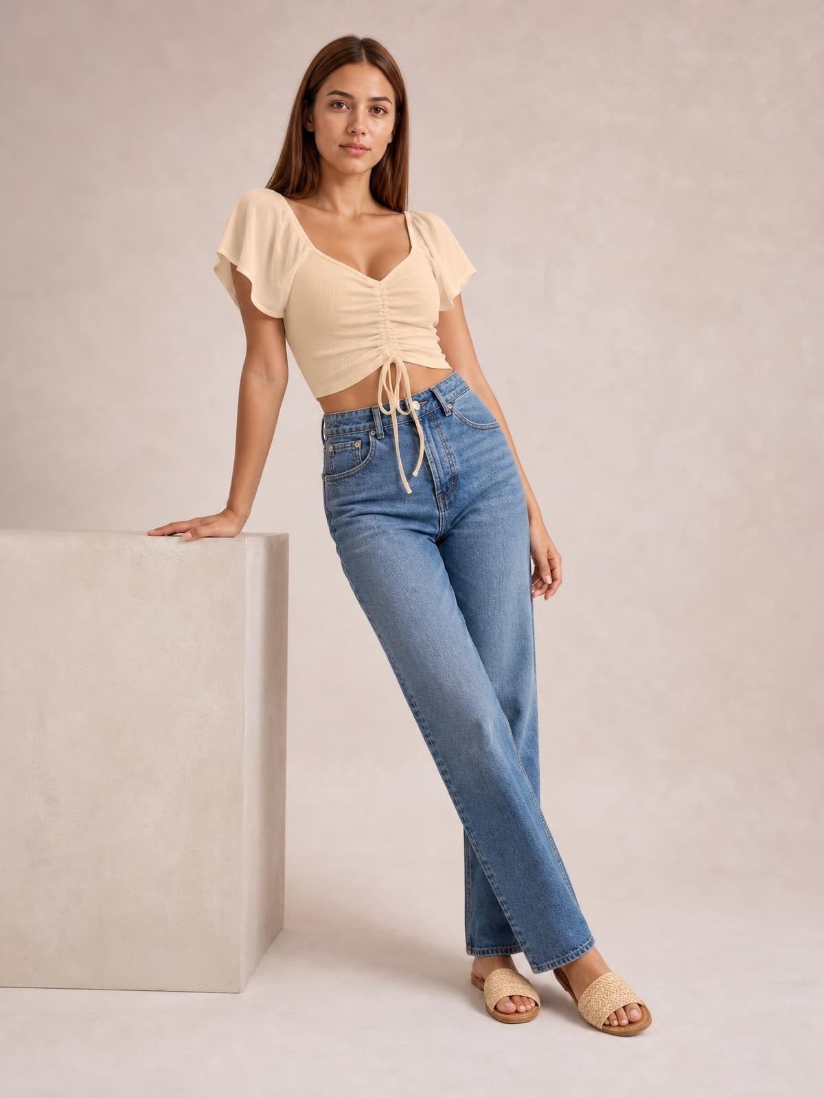 Model leans with one hand supported, wearing a cream drawstring short sleeve crop top with a V-neckline, ruched center, and cropped silhouette.