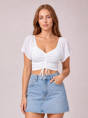 Model wearing a white drawstring short sleeve crop top with a ruched center, sweetheart neckline, and fitted silhouette, paired with a denim mini skirt.