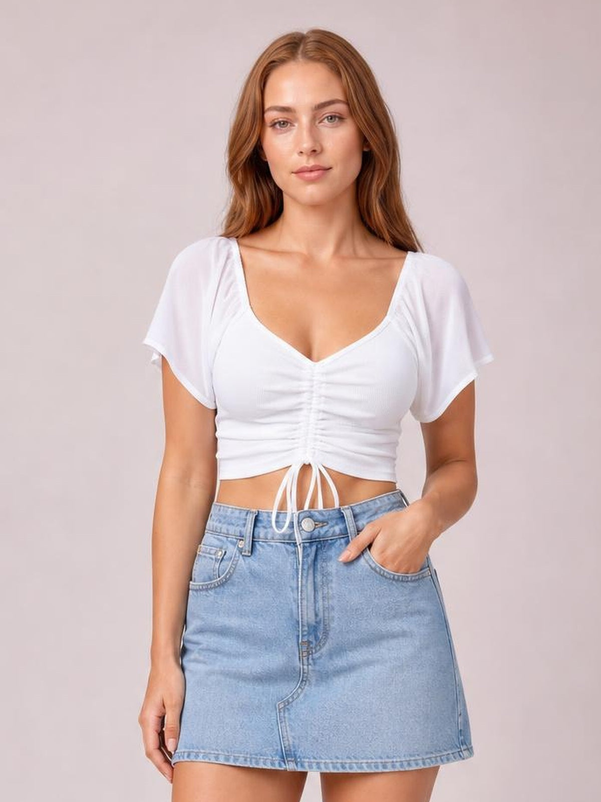 Model wearing a white drawstring short sleeve crop top with a ruched center, sweetheart neckline, and fitted silhouette, paired with a denim mini skirt.