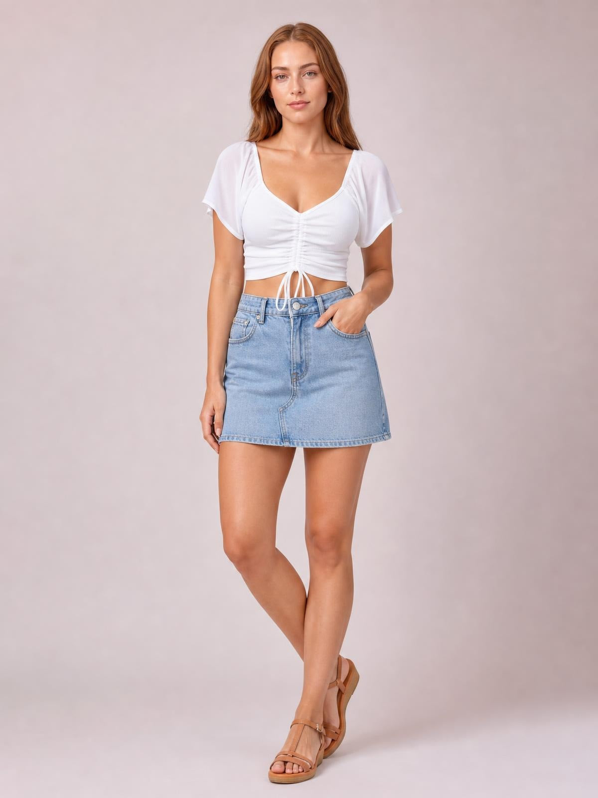 A model wears a white drawstring short sleeve crop top with a V-neck, ruched front, and tie hem, posing with crossed legs and one hand in her pocket.