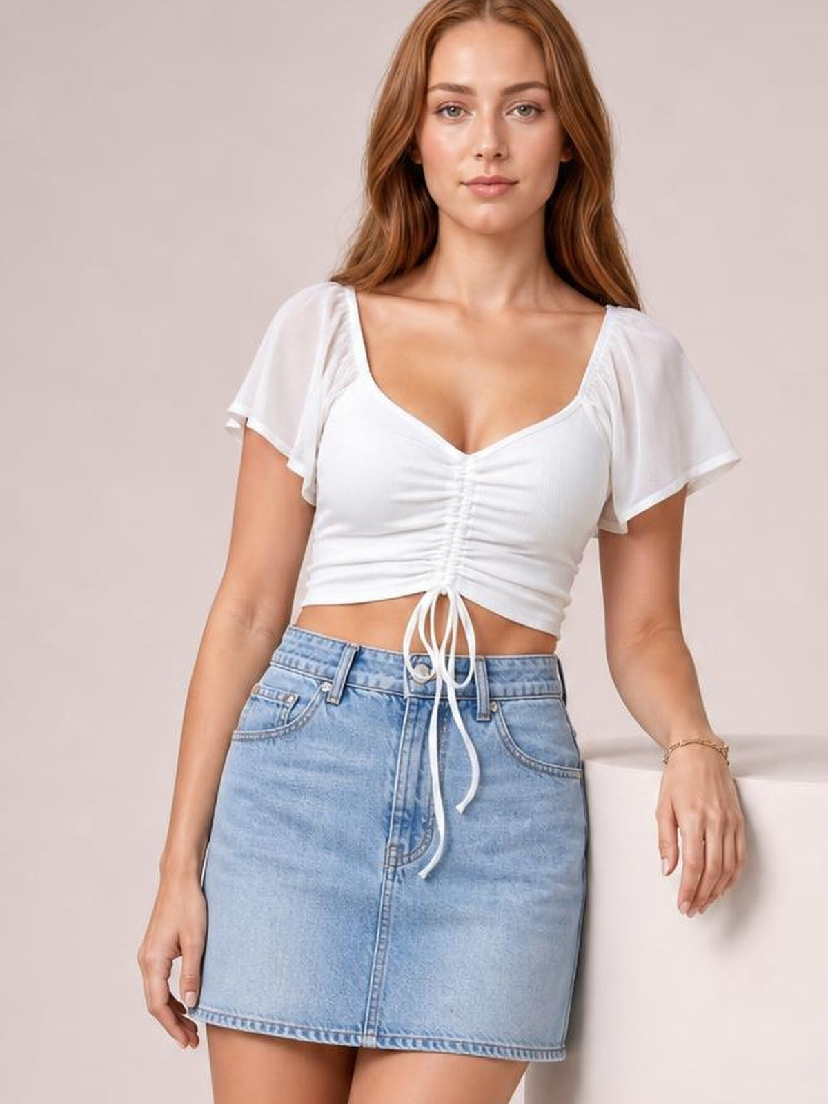 Model in a white short-sleeve crop top with a ruched drawstring front and tie, fitted silhouette, and moderate stretch, paired with a denim skirt.