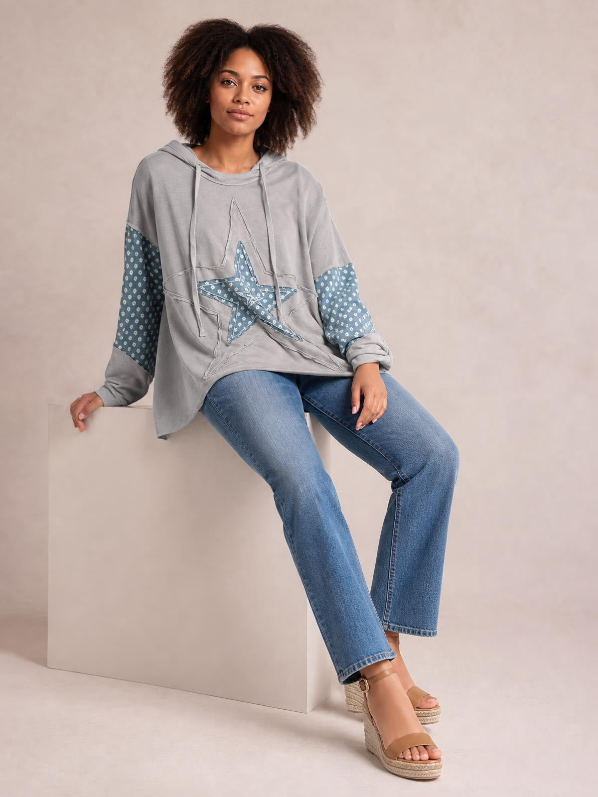 A seated model wears a loose gray drawstring star long sleeve hoodie with blue patterned sleeves, an oversized silhouette, and layered star appliqué detailing.