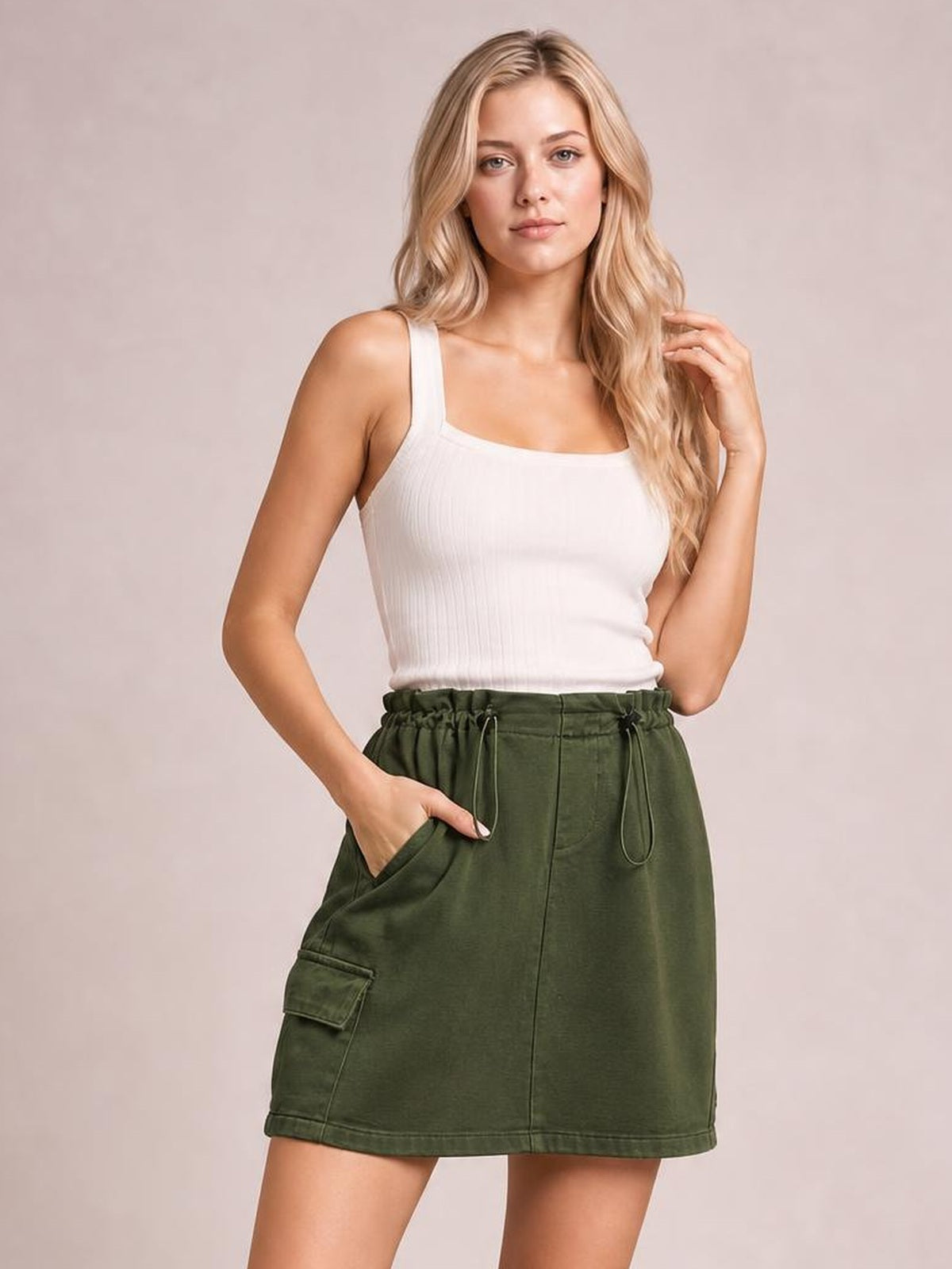 A model poses in an olive green denim mini skirt with a straight hem, drawstring waist, visible side pockets, and a flap cargo pocket in a relaxed silhouette.