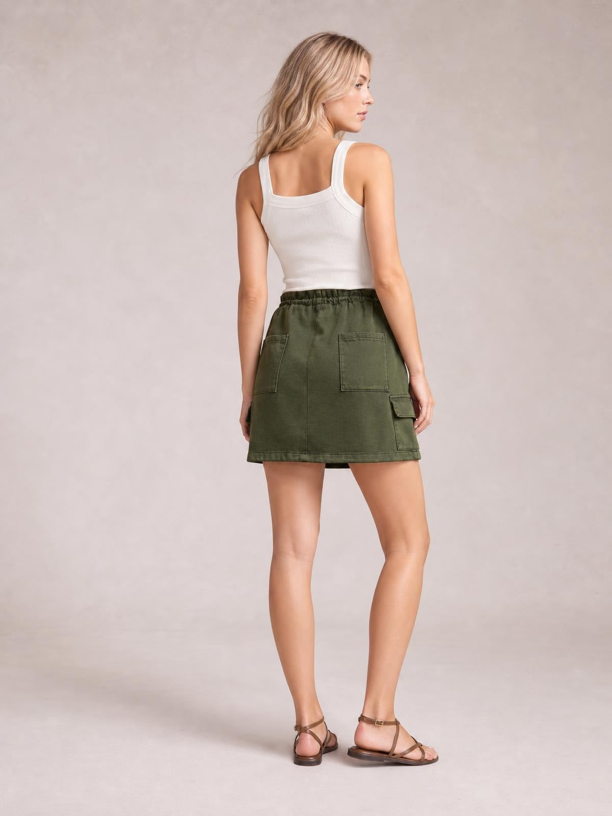 A model poses from the back wearing an olive drawstring mini skirt with a straight hem, utility-style patch pockets, and a relaxed silhouette.