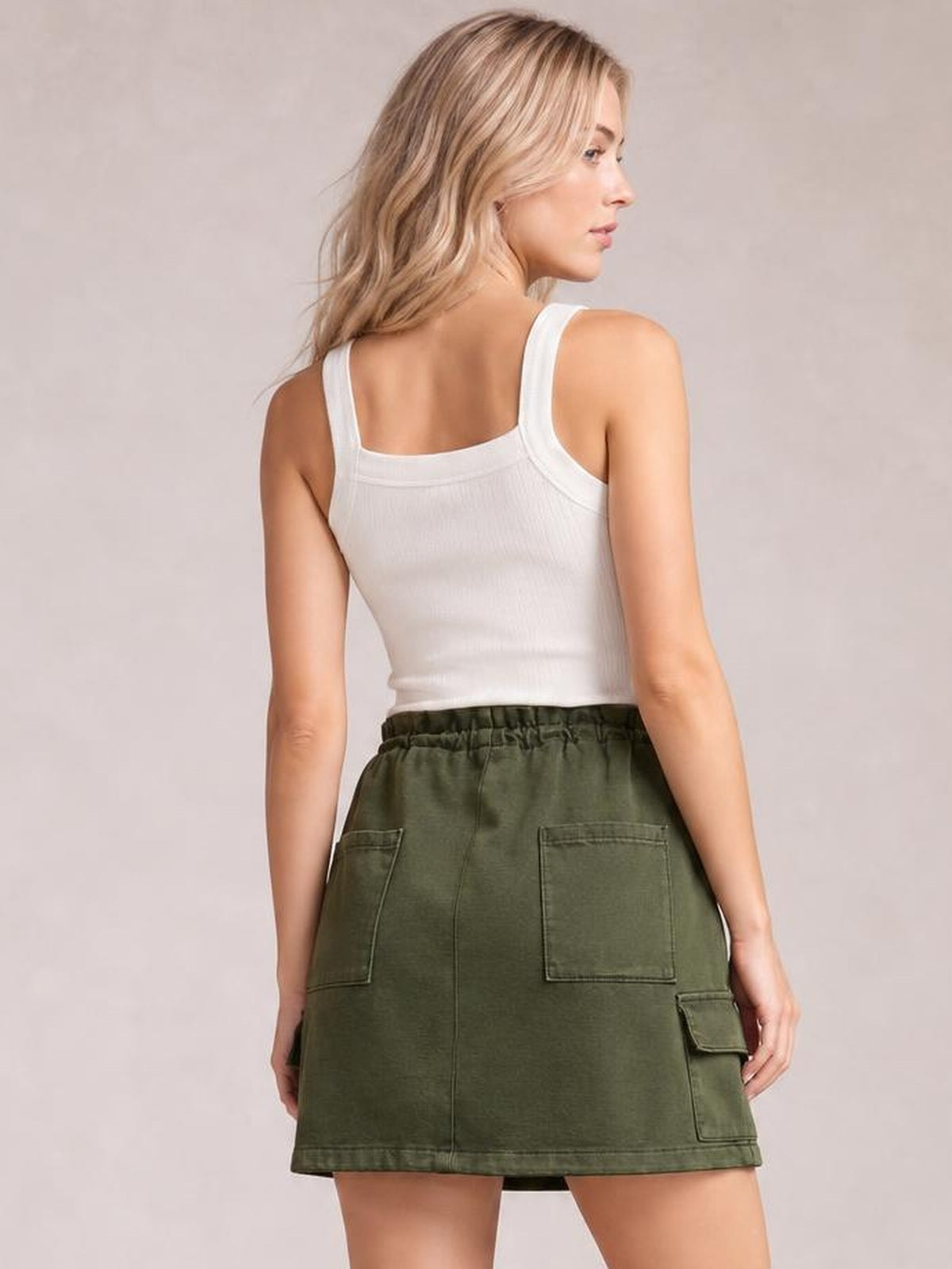 A model poses turned away in an olive green denim mini skirt with a drawstring waist, straight hem, and two large back patch pockets.