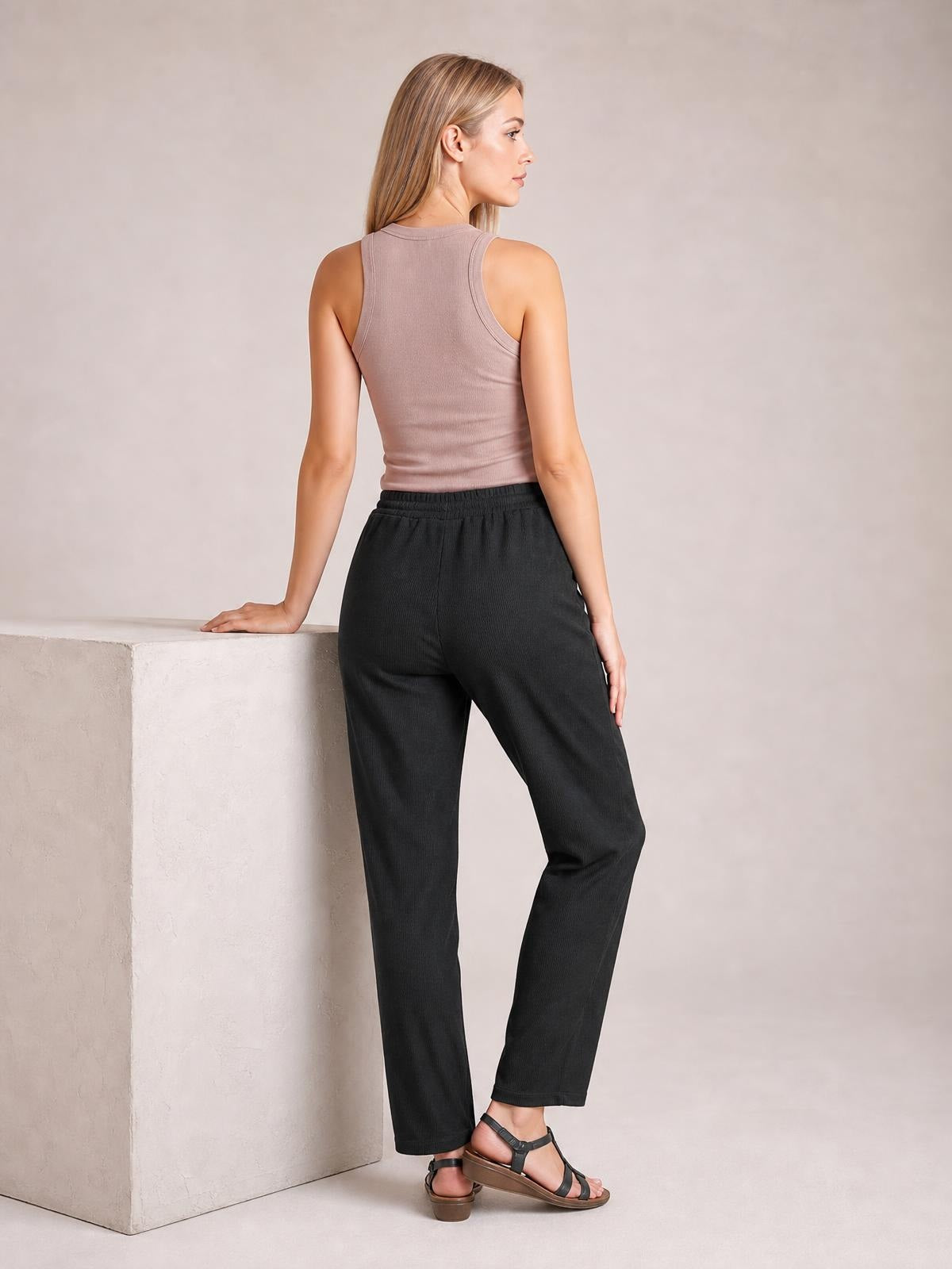 Model poses in black drawstring straight pants with a relaxed silhouette, elasticized waist, and side pockets, shown from the back.