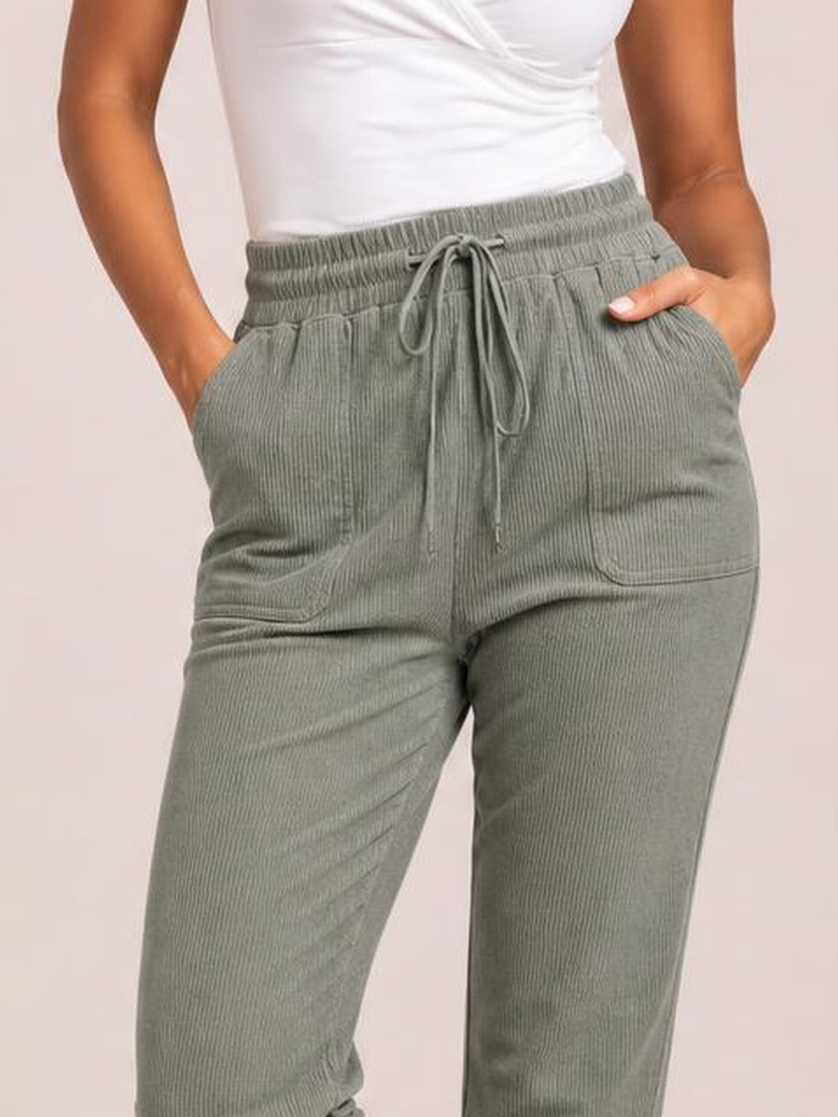 Model wearing gray-green straight pants with an elastic drawstring waist, side pockets, and a subtly textured construction, standing with hands tucked inside.