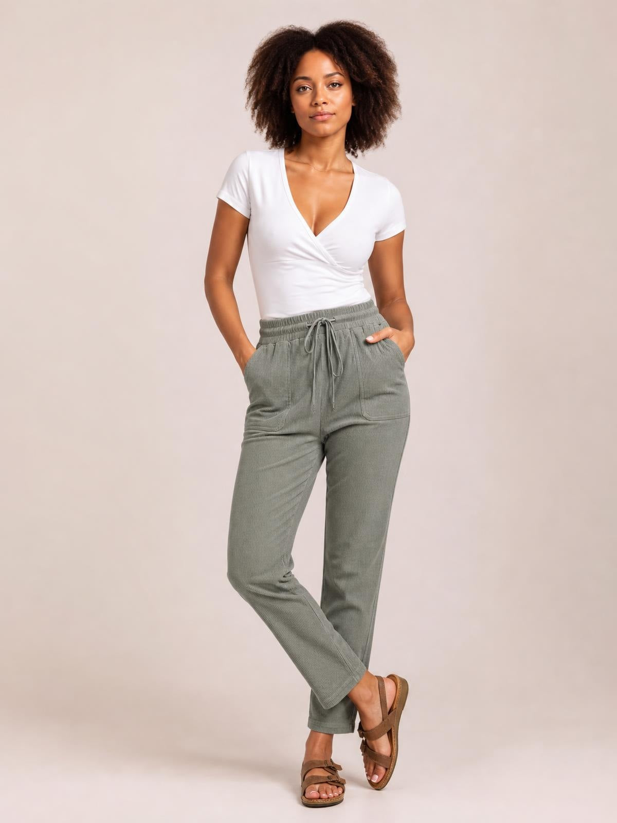 Model wears sage-gray straight-leg pants with a drawstring waist and roomy front pockets, posing with hands tucked in and one leg crossed.