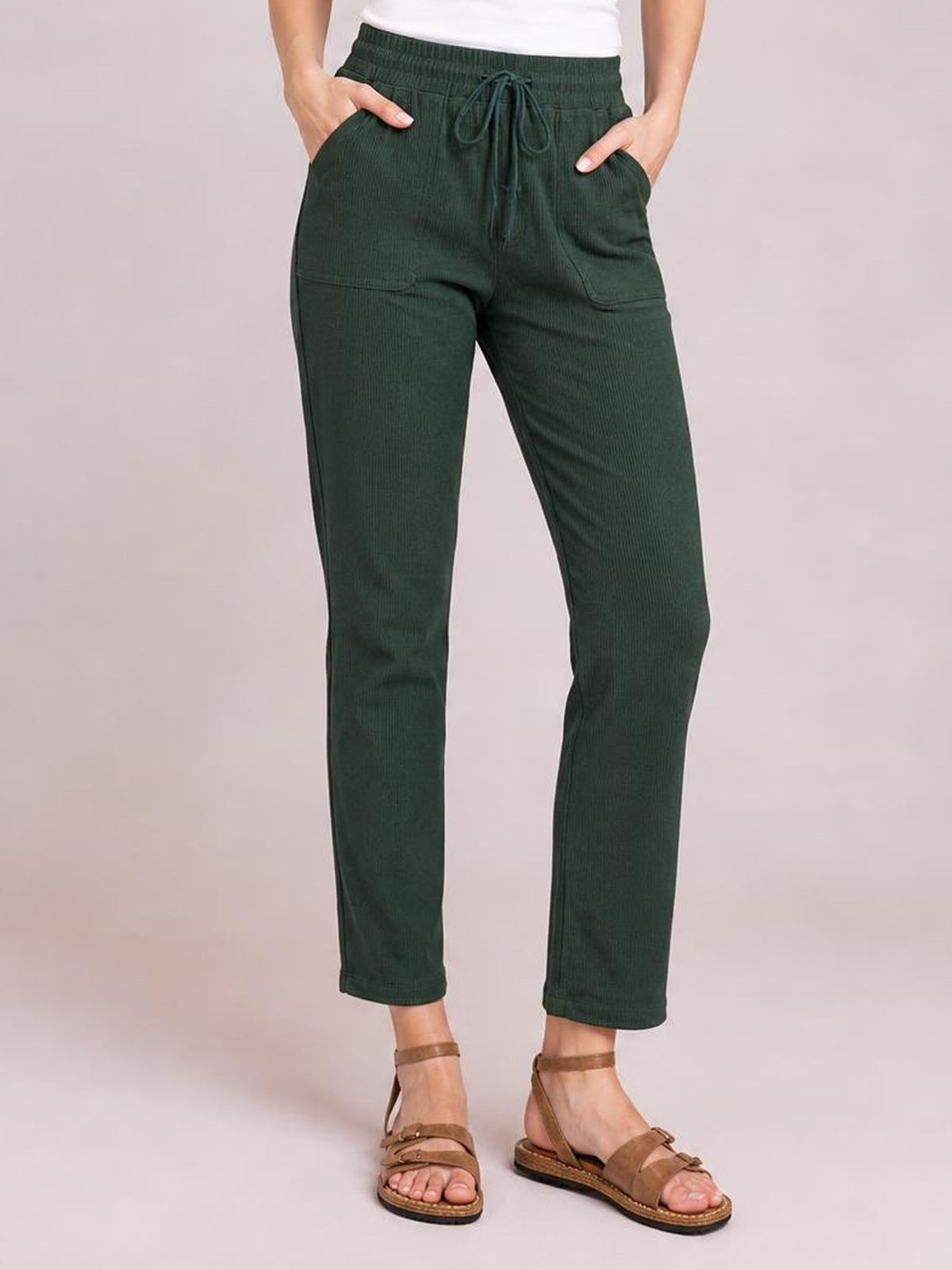 Model wears dark green straight-leg pants with a drawstring waistband and front pockets, standing with hands tucked in the pockets and ankle-length hems.