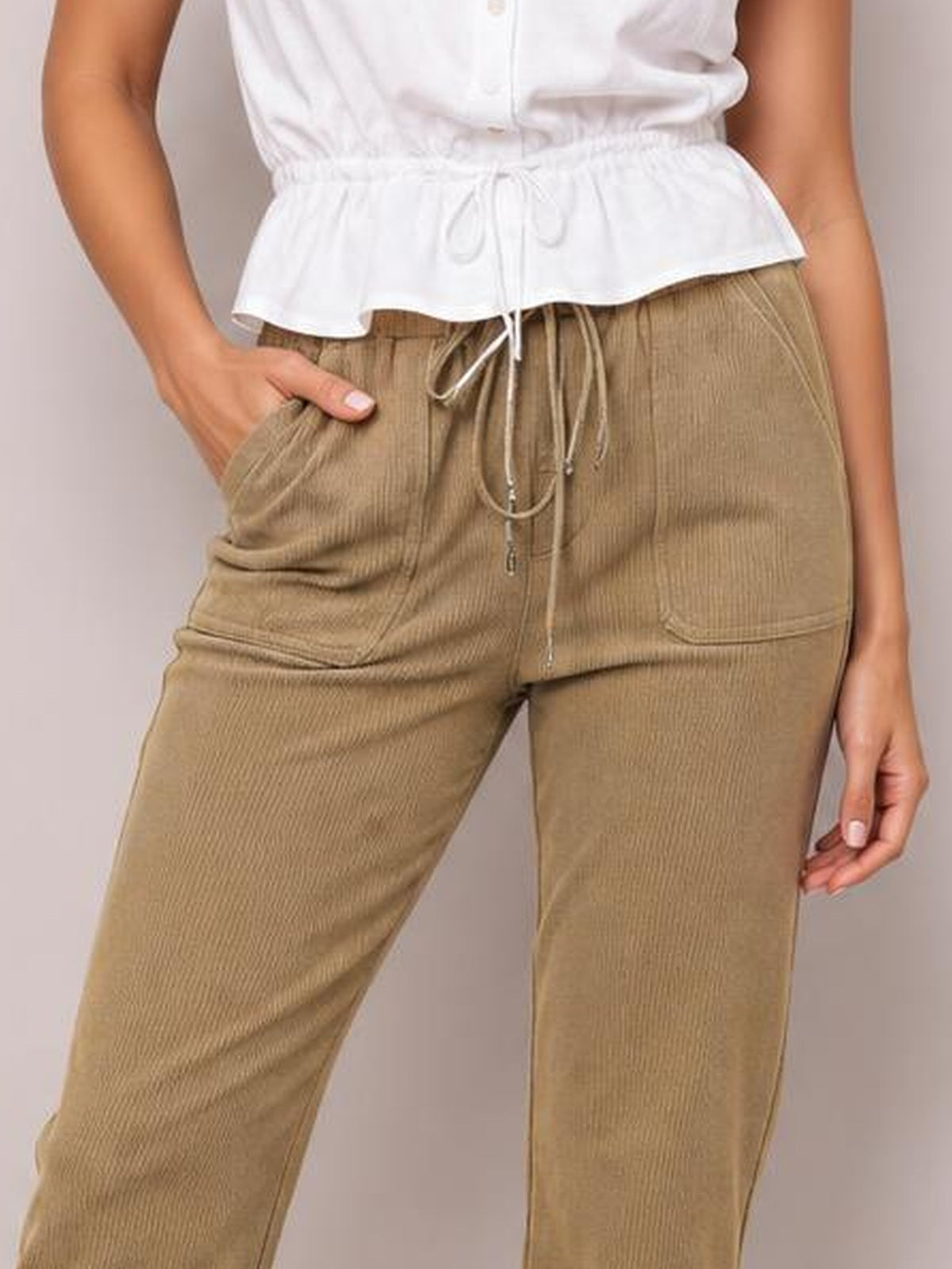 A model wears tan straight-leg pants with a drawstring waistband, visible front pockets, and textured fabric, posing with one hand tucked into a pocket.