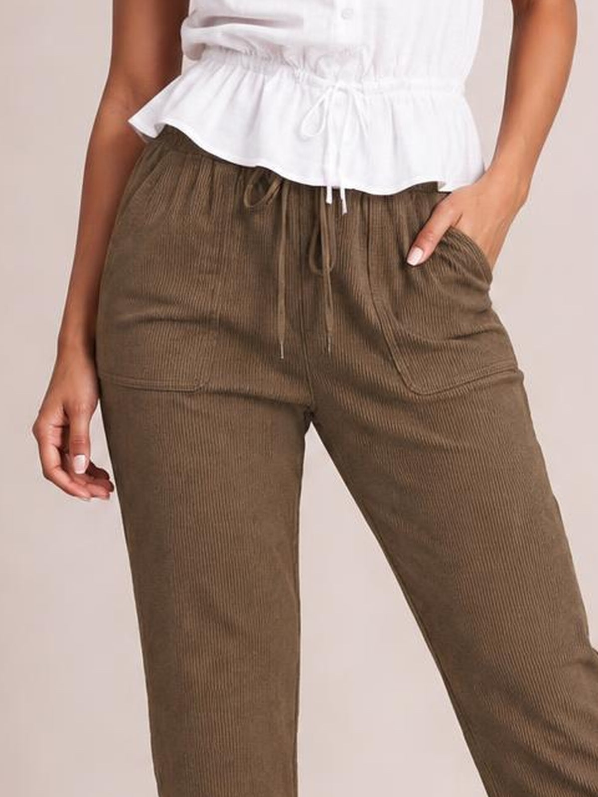 A model wears brown straight-leg pants with an elastic drawstring waist, vertical ribbed texture, and roomy front pockets, posing with one hand tucked inside.