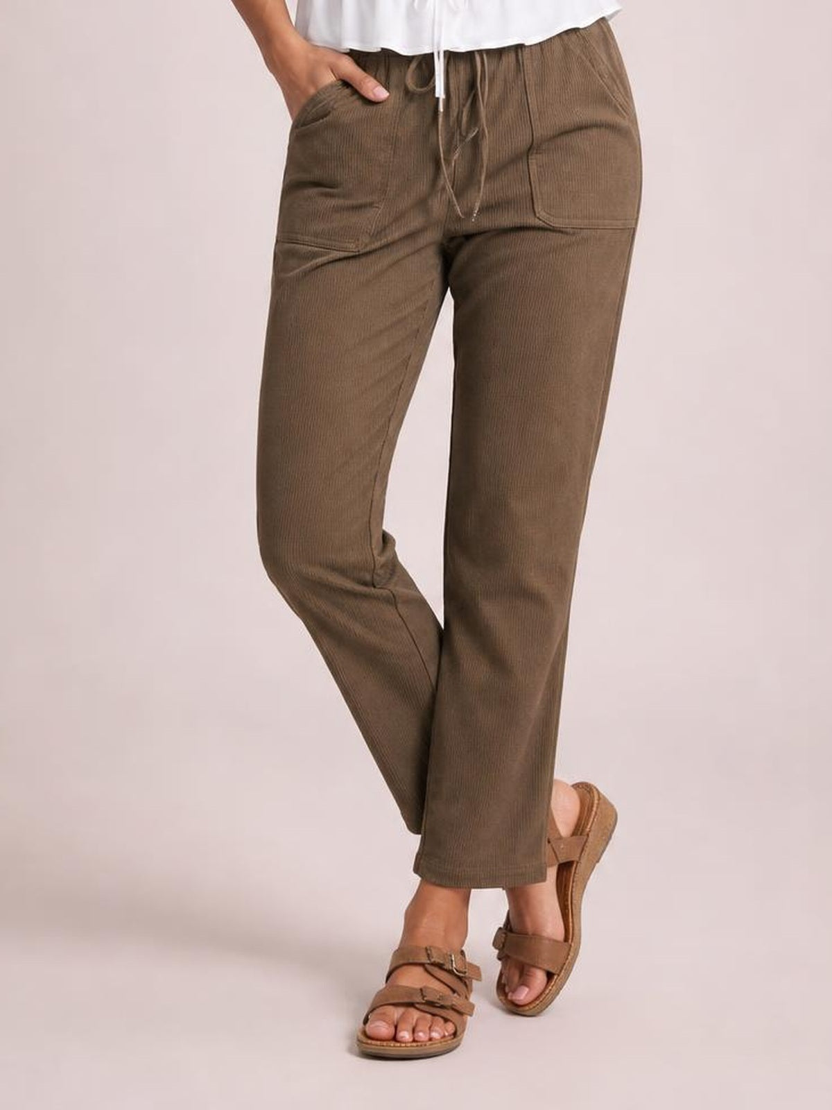 Model wears brown drawstring straight pants with visible front pockets and stitched panel details, posing with one hand in a pocket and crossed legs.