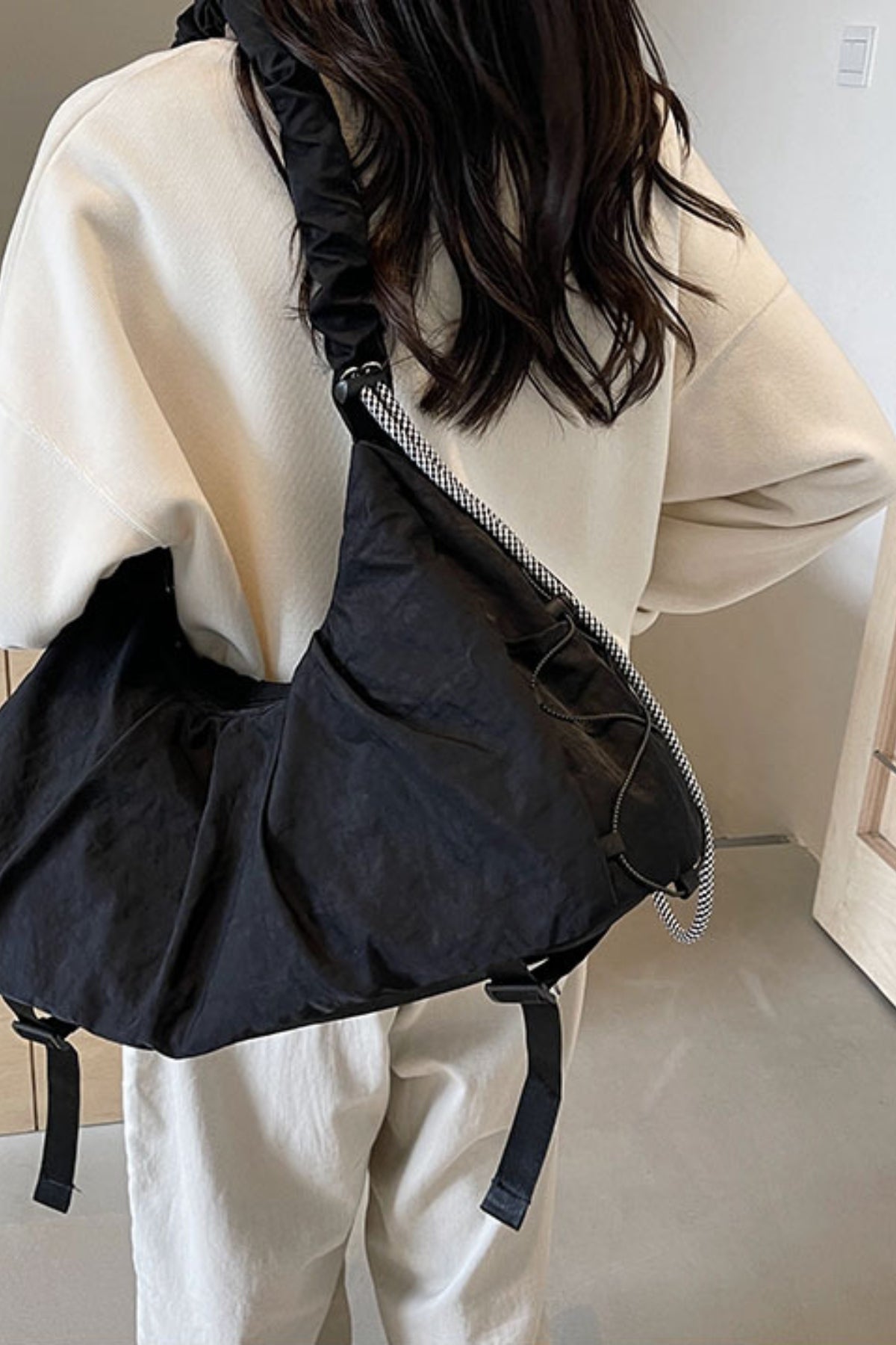 A model stands in a cream top and white pants, wearing a large black slouchy crossbody bag with a ruched drawstring strap across her back.