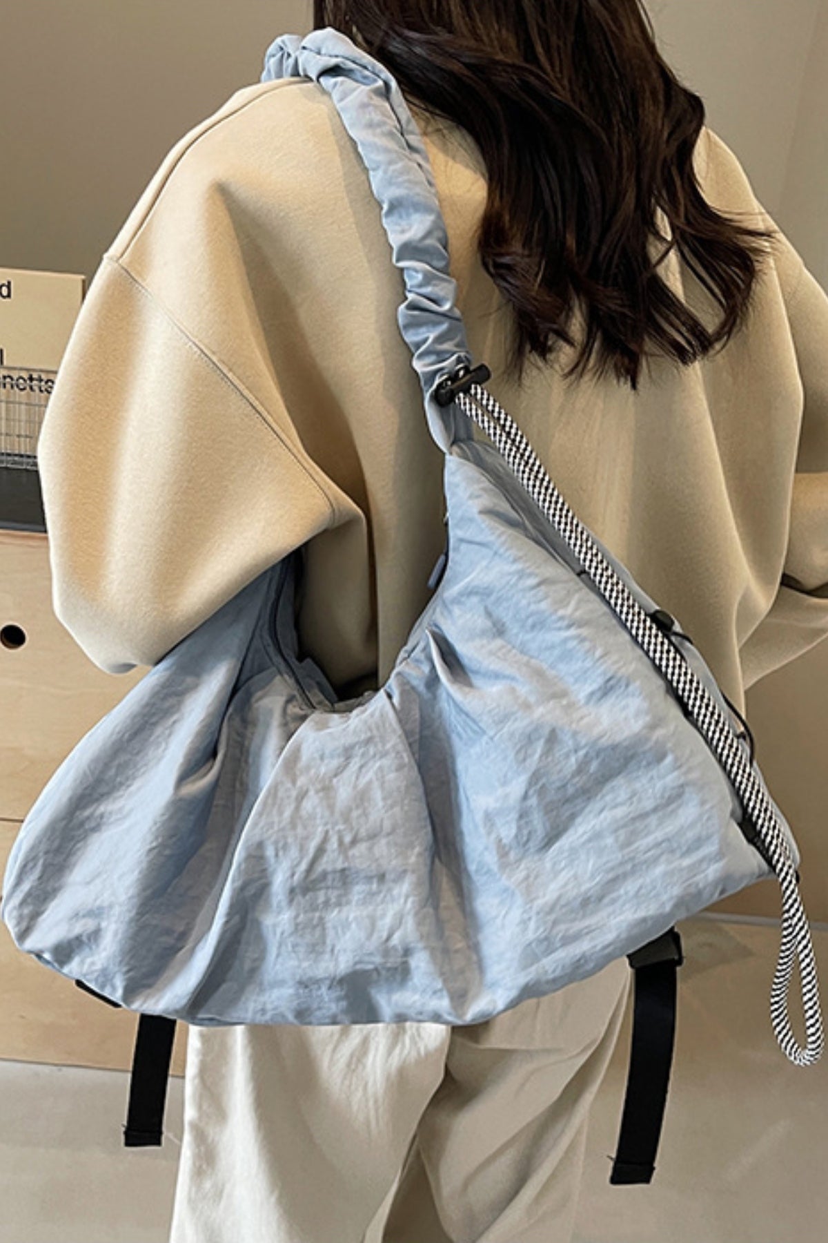 A model wears a large pale blue ruched crossbody bag across the back, featuring a slouchy silhouette and gathered drawstring strap over a beige oversized top.