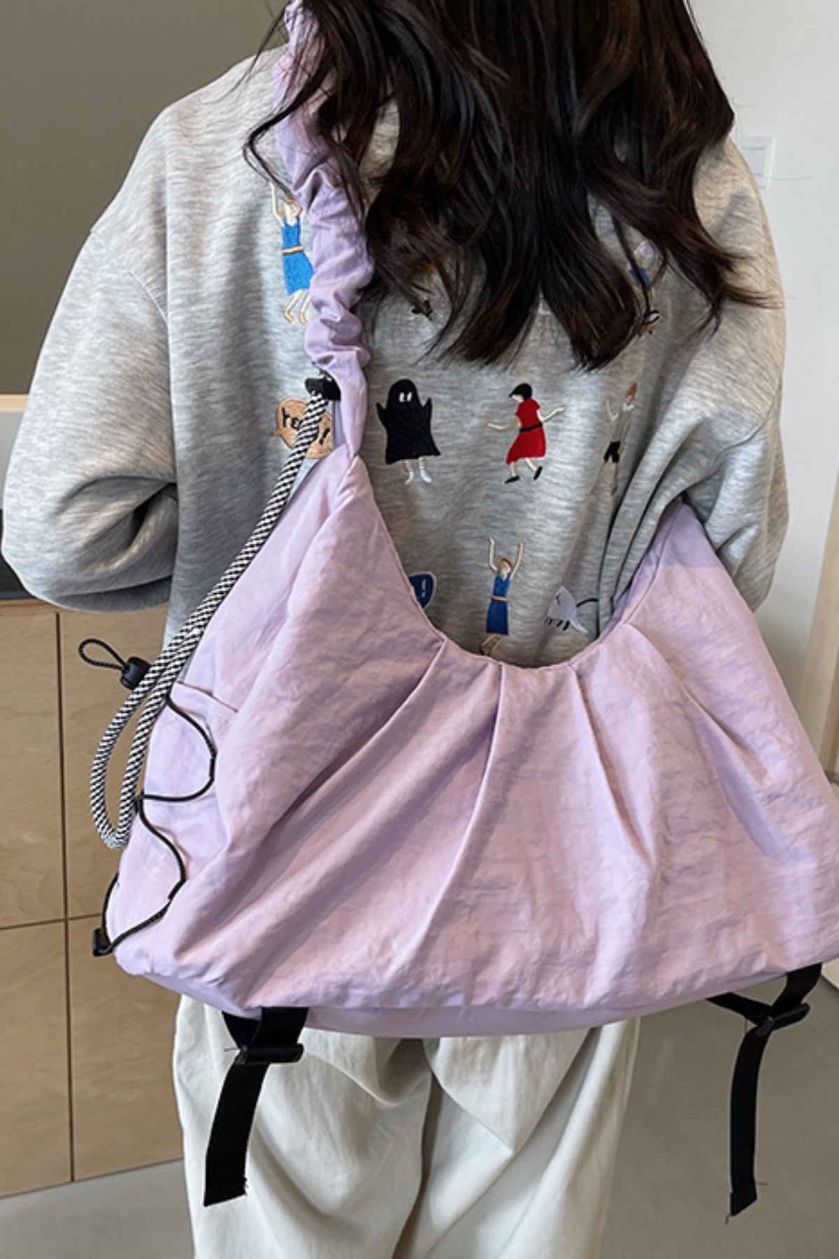 Model wears a large lavender ruched crossbody bag with a slouchy silhouette, gathered construction, and black drawstring strap over a gray sweatshirt.